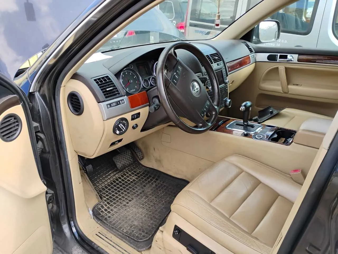 Wnętrze samochodu marki Volkswagen z kierownicą po lewej stronie. Deska rozdzielcza w kolorze beżowym z brązowymi akcentami drewnianymi. Widoczne są wskaźniki prędkości, obrotomierz, klimatyzacja, przyciski sterujące oraz automatyczna skrzynia biegów. Drzwi kierowcy są otwarte, pokazując panel drzwiowy z głośnikiem i sterownikami. Na podłodze czarne maty z siatkowym wzorem. Siedzenia tapicerowane beżową skórą. W tle przez przednią szybę widoczne są inne pojazdy i fragment budynku.