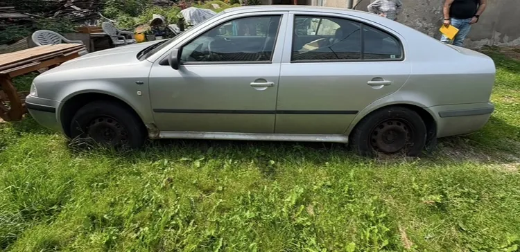 Srebrny samochód osobowy typu sedan zaparkowany na trawiastym terenie. Pojazd jest pokazany z profilu od strony kierowcy. W tle widoczne są elementy podwórka, w tym drewniany stół, krzesła ogrodowe oraz fragmenty budynków.