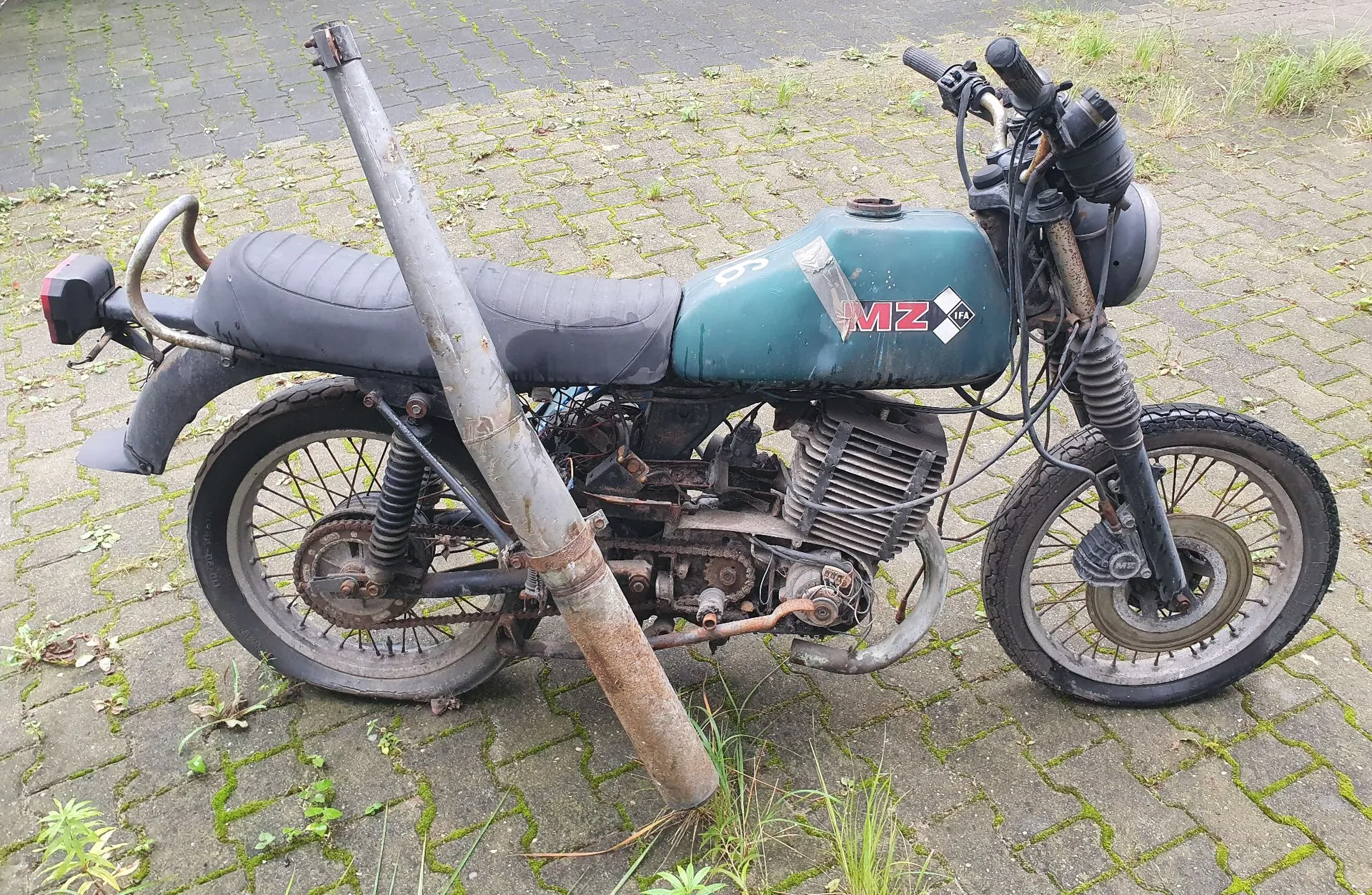Stary, zniszczony motocykl marki MZ w kolorze ciemnozielonym, zaparkowany na kostce brukowej porośniętej trawą. Motocykl ma czarne siedzenie, widoczne logo 'MZ' na baku oraz zardzewiałe elementy silnika i ramy. Przed motocyklem, oparty o jego ramę, leży długi, zardzewiały metalowy rurka lub pręt.