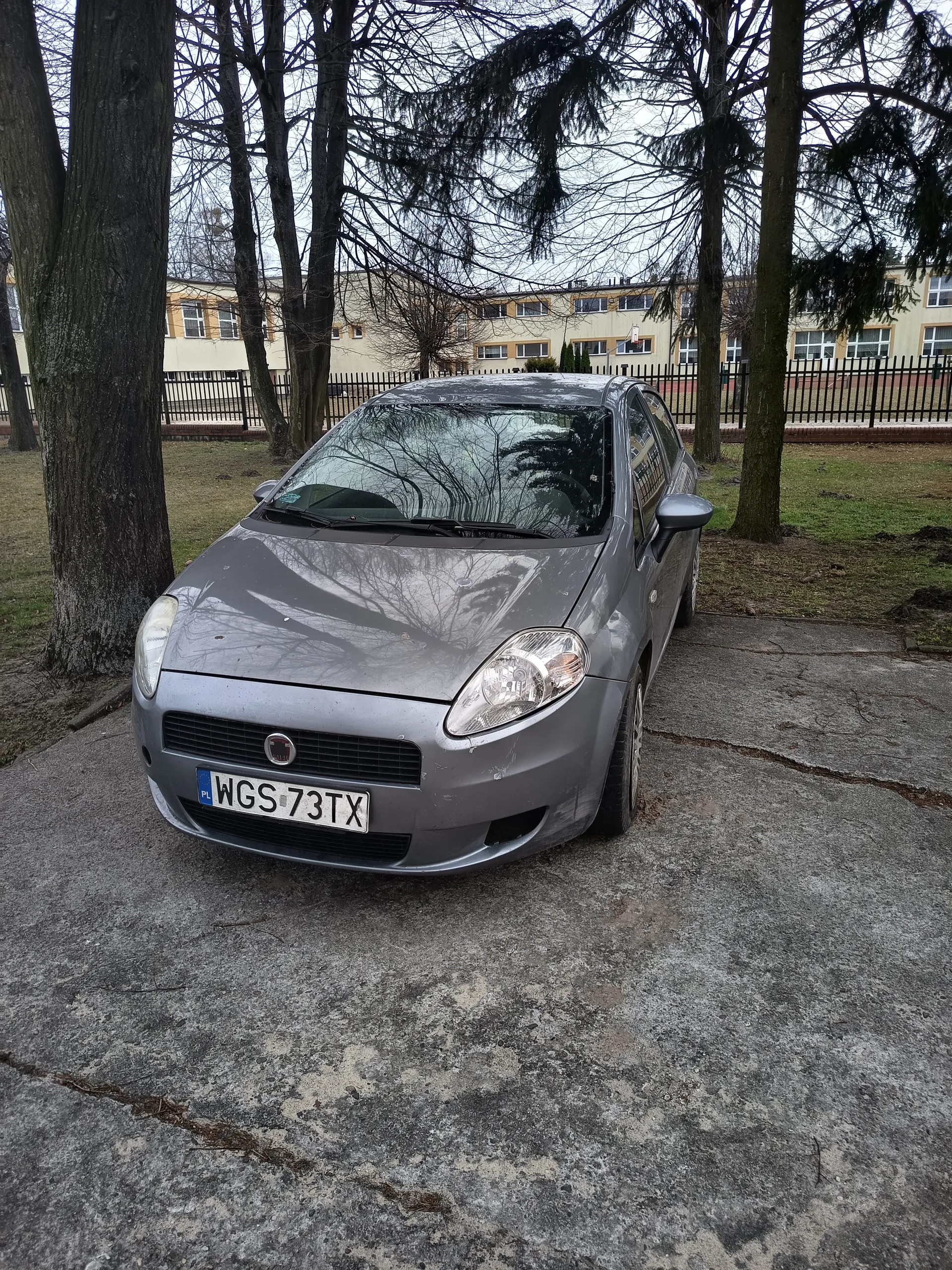 Srebrny samochód marki Fiat zaparkowany na betonowej powierzchni. Widoczna tablica rejestracyjna z polskim kodem PL i numerem WGS 73TX. W tle drzewa bez liści, czarny metalowy płot oraz wielopiętrowy budynek o żółto-białej elewacji z oknami. Niebo jest zachmurzone, a na ziemi znajdują się fragmenty trawy i suche gałęzie.