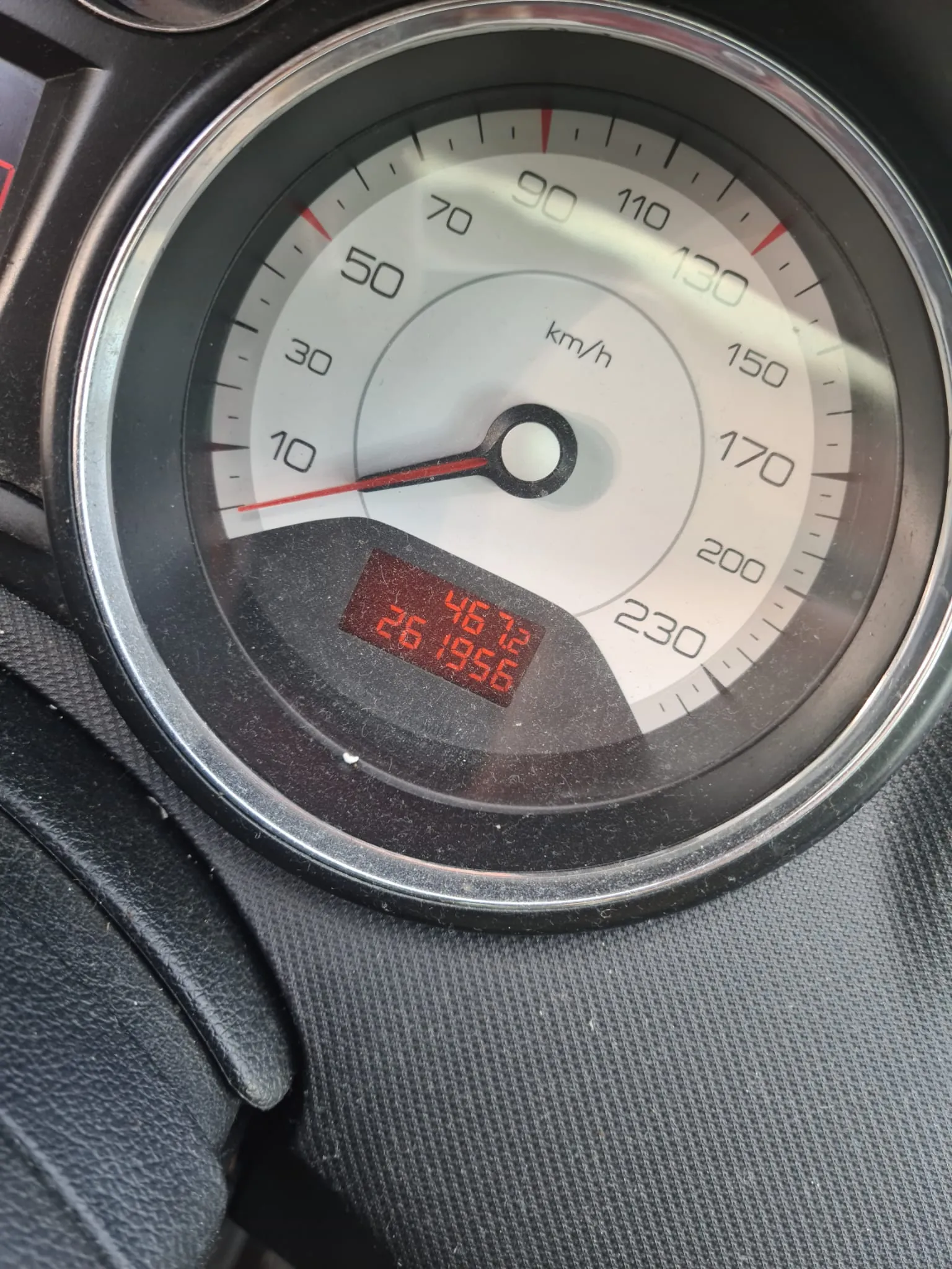Zbliżenie na samochodowy wskaźnik prędkości z białą tarczą i czarnymi cyframi od 0 do 230 km/h. Czerwona strzałka wskazuje prędkość bliską 0 km/h. W środku tarczy widoczny napis 'km/h'. Poniżej znajduje się czerwony wyświetlacz cyfrowy z liczbami '46.2' i '26 1956'. Tło stanowi czarna, teksturowana deska rozdzielcza.