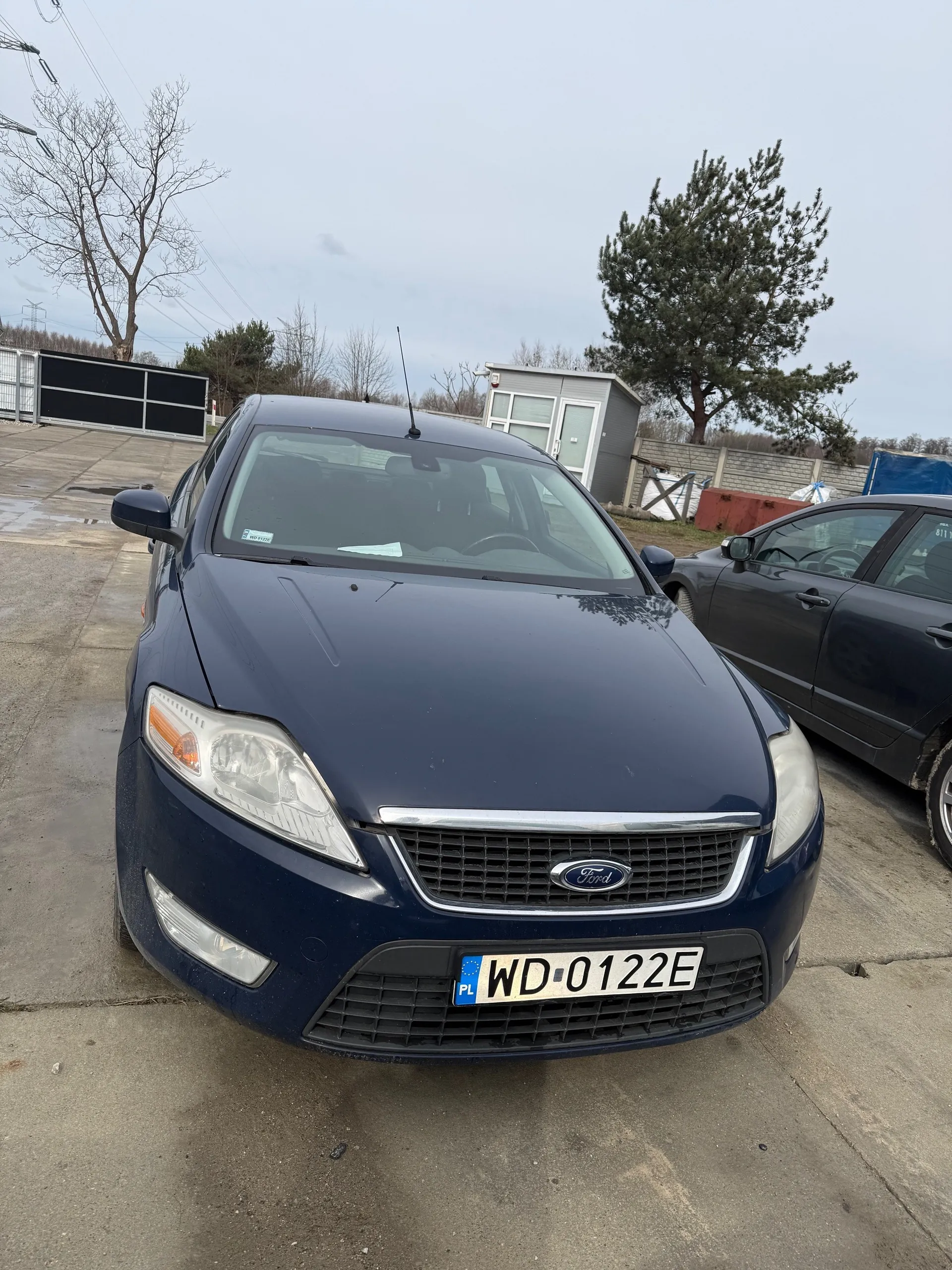 Ciemnoniebieski samochód marki Ford z tablicą rejestracyjną PL WD-0122E zaparkowany na betonowej powierzchni. W tle drugi ciemnoszary samochód, szara budka z oknami, drzewa bez liści, iglaste drzewo, ogrodzenie, czerwony kontener i linie energetyczne. Niebo jest pochmurne.