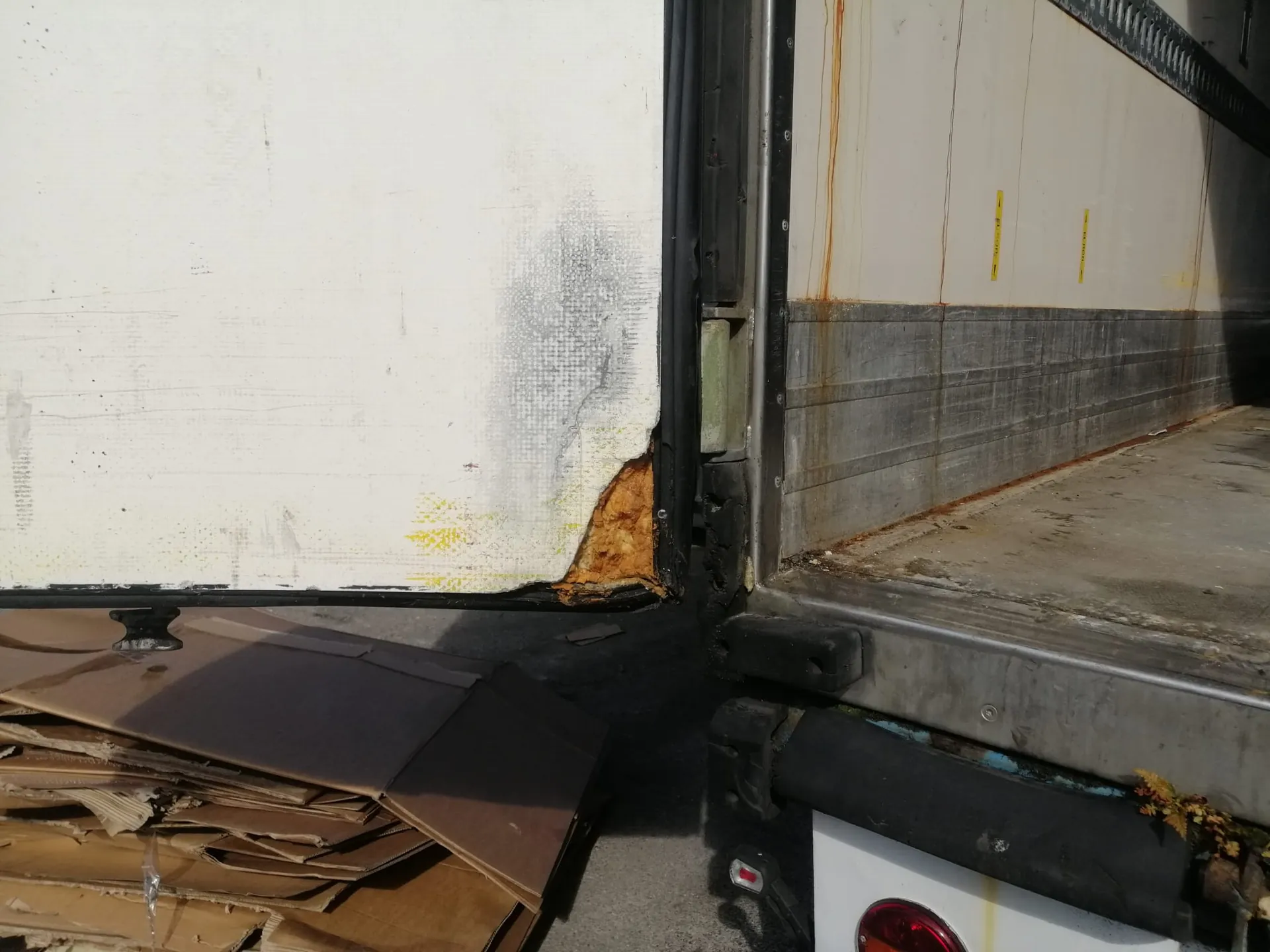 Zbliżenie na uszkodzenie w dolnej części białej ściany naczepy pojazdu transportowego. W narożniku widoczny jest ubytek materiału, odsłaniający brązową izolację termiczną. Powierzchnia wokół uszkodzenia jest zabrudzona i widać na niej zacieki. Obok znajduje się otwarta przestrzeń ładunkowa z metalową podłogą oraz stosy brązowych kartonów na ziemi pod pojazdem.