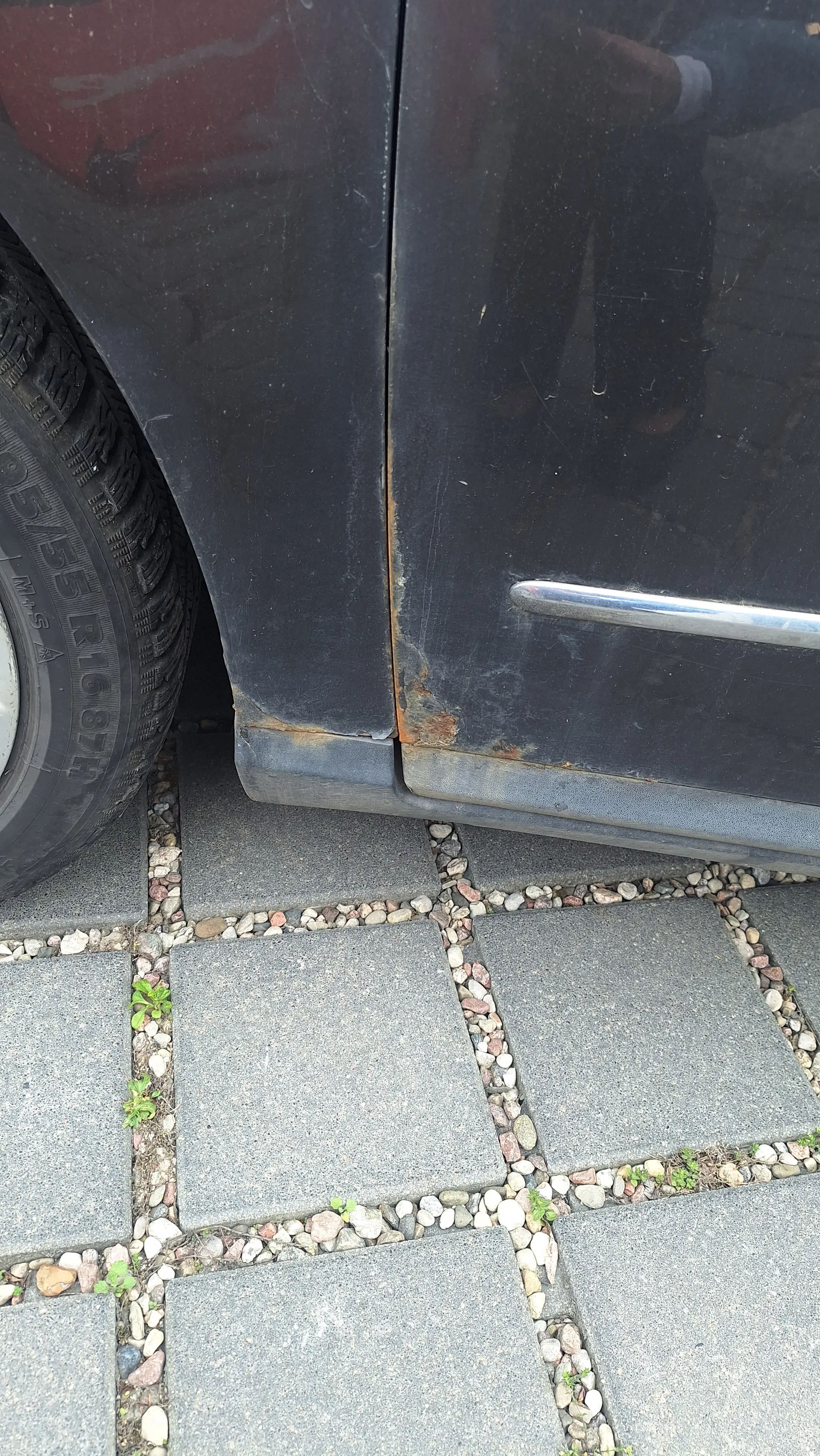 Zbliżenie na tylną część czarnego samochodu z widoczną rdzą na dolnej krawędzi karoserii. Częściowo widoczna opona z napisem 205/55 R16 87H oraz M+S. Podłoga wyłożona kwadratowymi płytami betonowymi oddzielonymi małymi kamieniami, między którymi rosną zielone rośliny.