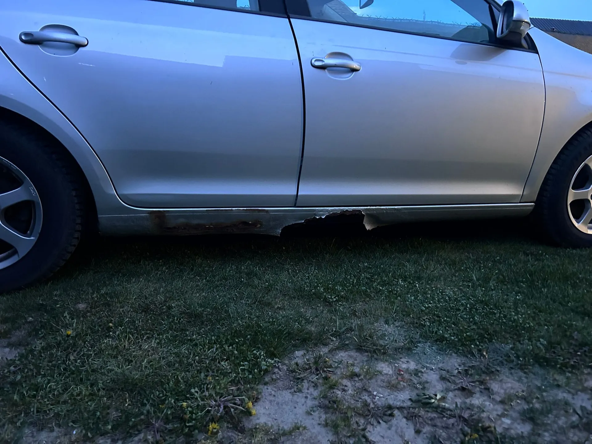 Zbliżenie na prawy bok srebrnego samochodu, pokazujące dolną krawędź drzwi i progu. W dolnej części nadwozia, wzdłuż progu, widoczne są znaczne uszkodzenia w postaci rdzy oraz ubytków w blachu (przerdzewienia). Samochód stoi na trawiastym podłożu.
