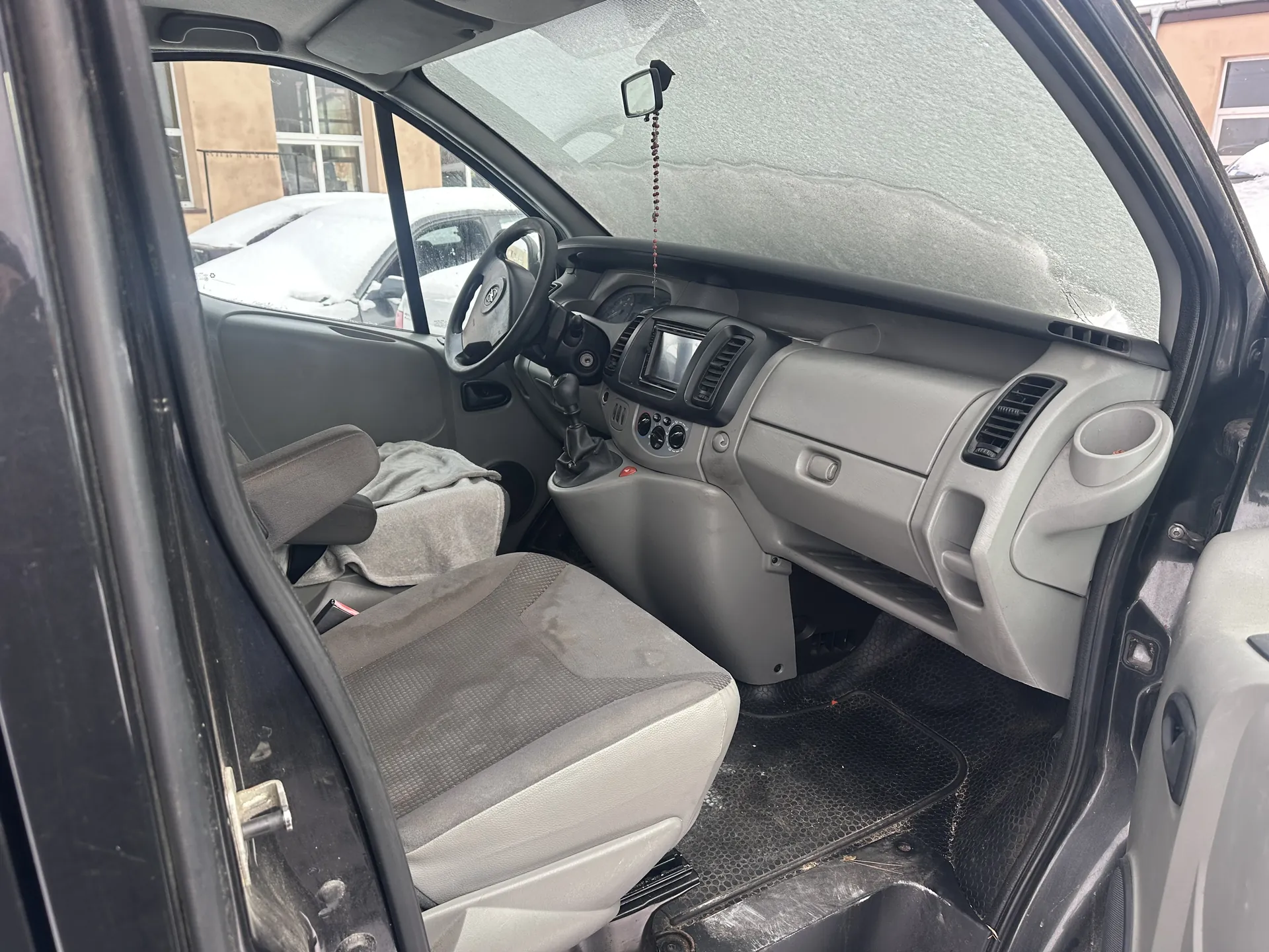 Wnętrze samochodu z otwartą drzwiami kierowcy. Widoczne są szare siedzenia, na siedzeniu pasażera leży szary ręcznik. Deska rozdzielcza w kolorze szarym z małym ekranem, klimatyzacją i przyciskami. Kierownica po lewej stronie, lusterko wsteczne z wiszącym ozdobnym elementem. Przez przednią szybę widać zaparkowane samochody pokryte śniegiem oraz budynki. Podłoga wyłożona czarną gumową wykładziną.