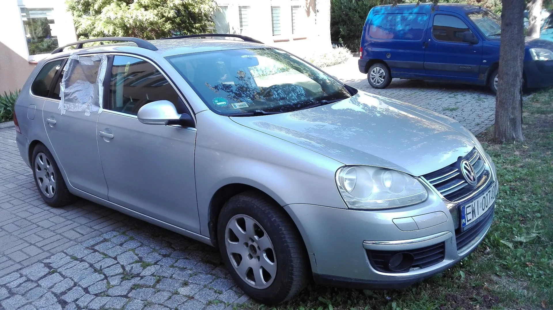 Srebrny samochód osobowy marki Volkswagen w nadwoziu kombi, zaparkowany na kostce brukowej. Przednia lewa szyba pasażera jest zaklejona przezroczystą folią ochronną. W tle widoczny jest niebieski samochód dostawczy oraz fragment budynku i drzewa. Na samochodzie widoczna jest tablica rejestracyjna zaczynająca się od liter EW.