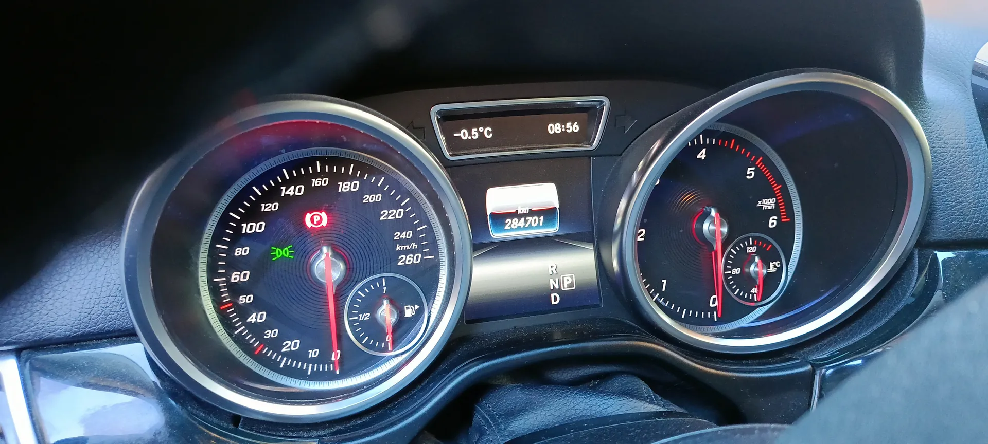 Zbliżenie na deski rozdzielcze samochodu. Po lewej stronie prędkościomierz z zakresem do 260 km/h, wskazówka na 0, czerwony symbol P (parking) i zielony symbol D (drive). W dolnej części prędkościomierza wskaźnik paliwa w połowie. Po prawej stronie tachometr z zakresem do 6 x1000 obr./min, wskazówka na 0. W centralnej części wyświetlacz cyfrowy z temperaturą -0.5°C, godziną 08:56 oraz przebiegiem 284701 km. Poniżej przebiegu widoczne oznaczenia biegów R, N, P, D z zaznaczonym P. Tło deski rozdzielczej czarne z metalowymi obramowaniami przyrządów.