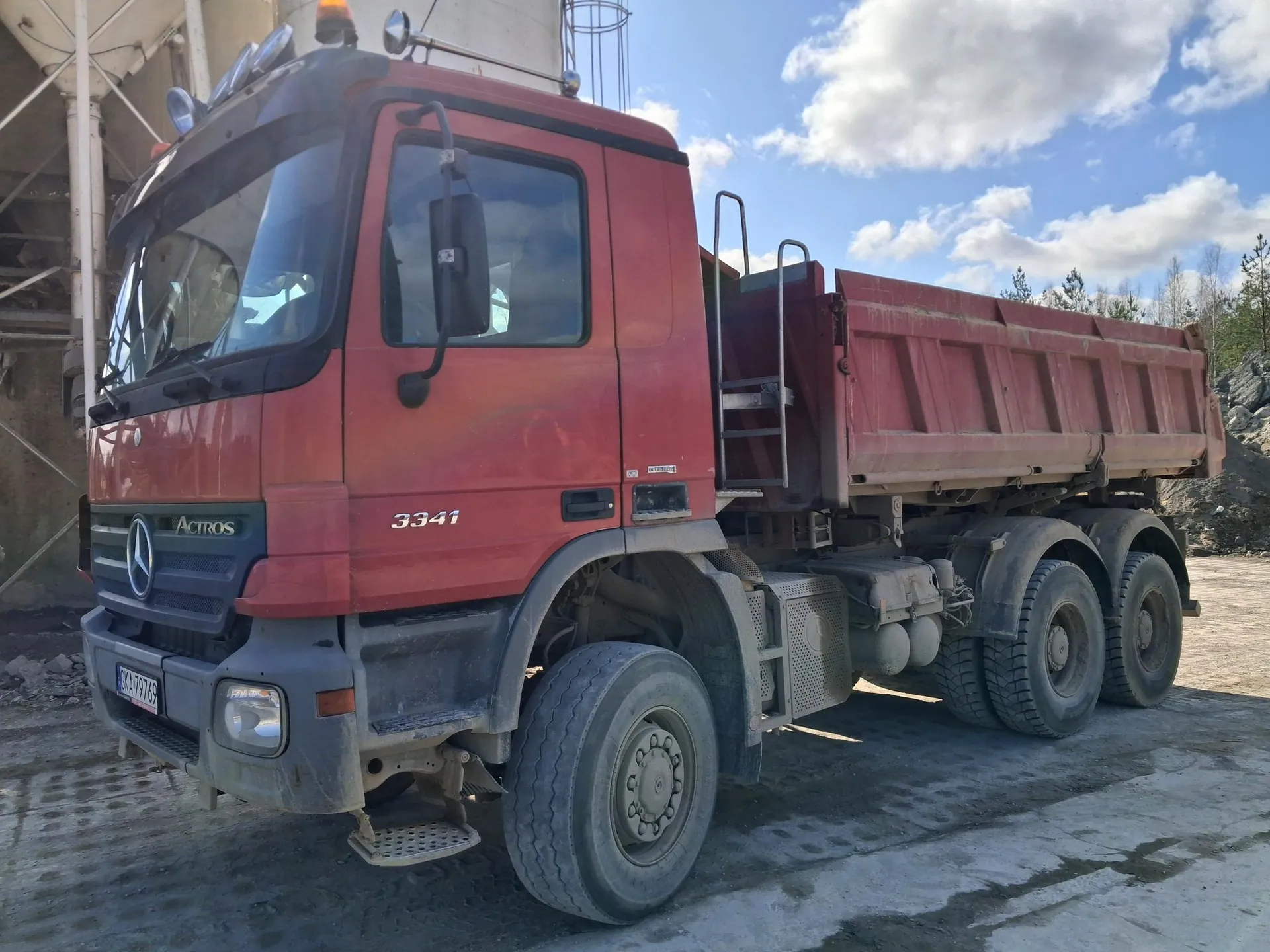Czerwona ciężarówka wywrotka marki Mercedes-Benz Actros 3341 z tablicą rejestracyjną BKA 79769. Pojazd stoi na betonowej powierzchni, w tle budowla przemysłowa, stos skał, drzewa i niebo z chmurami. Widoczne są duże opony, drabinka po boku kabiny oraz logo Mercedes-Benz na masce.