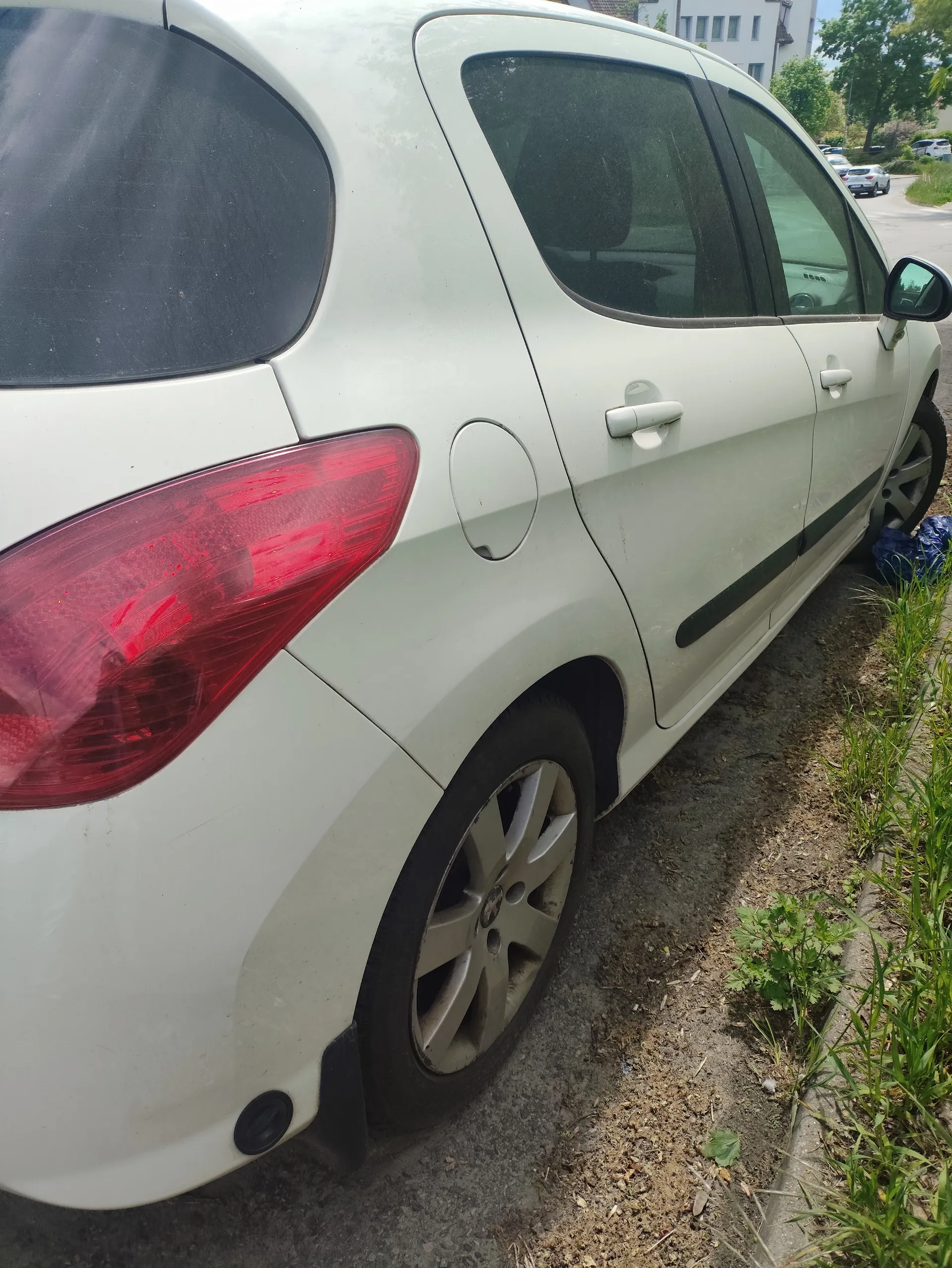 Biały samochód osobowy typu hatchback widziany z tyłu i boku. Czerwone światło tylnego reflektora, srebrne felgi, czarna listwa boczna. Samochód zaparkowany na nierównym podłożu obok trawy. W tle widoczne inne pojazdy, budynki i drzewa.
