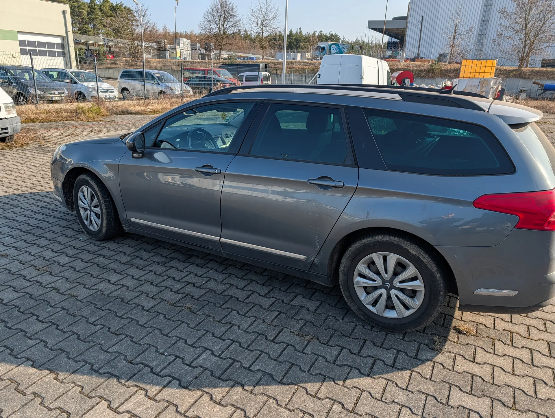 Szary samochód typu kombi zaparkowany na brukowanym placu. W tle widoczne są inne pojazdy, budynek przemysłowy z zielonym pasem, drzewa bez liści oraz ogrodzenie. Niebo jest jasne, a cień od samochodu wskazuje na słoneczny dzień. Brak czytelnego tekstu na obrazie.