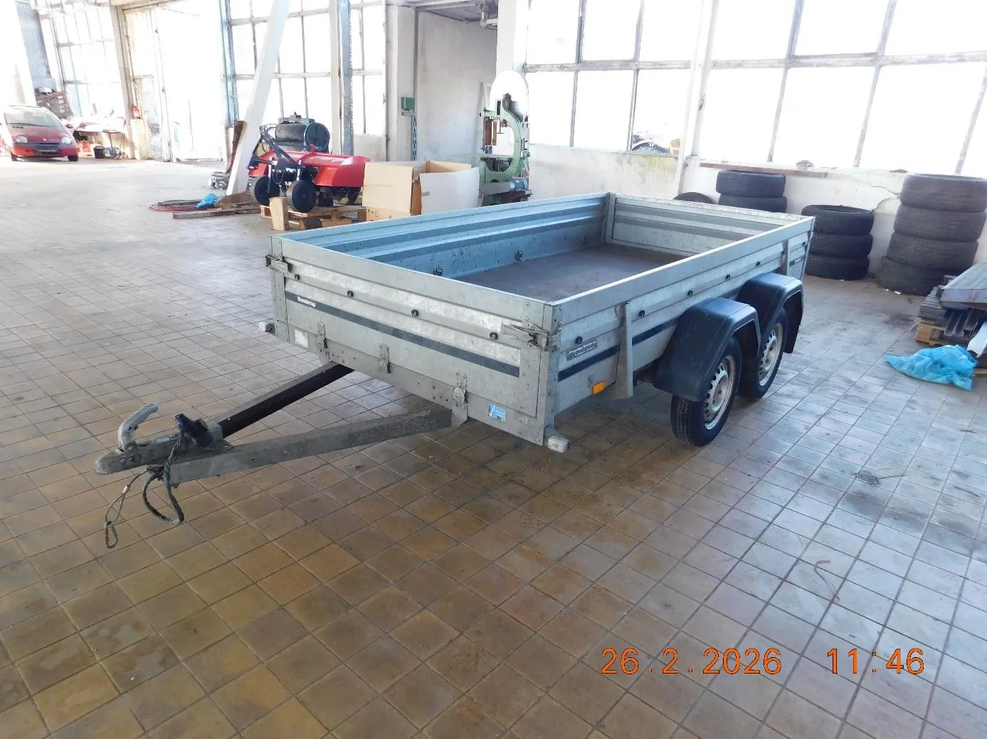 Metalowy przyczepa z dwoma osiami i hakem holowniczym umieszczona w przestronnym pomieszczeniu przemysłowym z podłogą z brązowych płytek. W tle widoczne są czerwony traktor, stosy opon, czerwony samochód, maszyny oraz duże okna. W prawym dolnym rogu obrazu znajduje się pomarańczowy znaczek z datą i godziną: 26.2.2026 11:46.