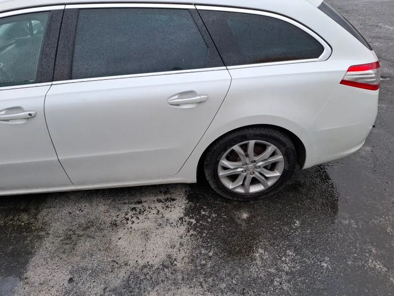 Biały samochód typu kombi zaparkowany na mokrym asfalcie. Widoczne są boczne drzwi, czarne przyciemniane szyby z kroplami deszczu, srebrne felgi oraz czerwono-białe światło tylnego. Asfalt pokryty jest plamami wilgoci i drobnymi kamieniami. Tło stanowi pusty parking.