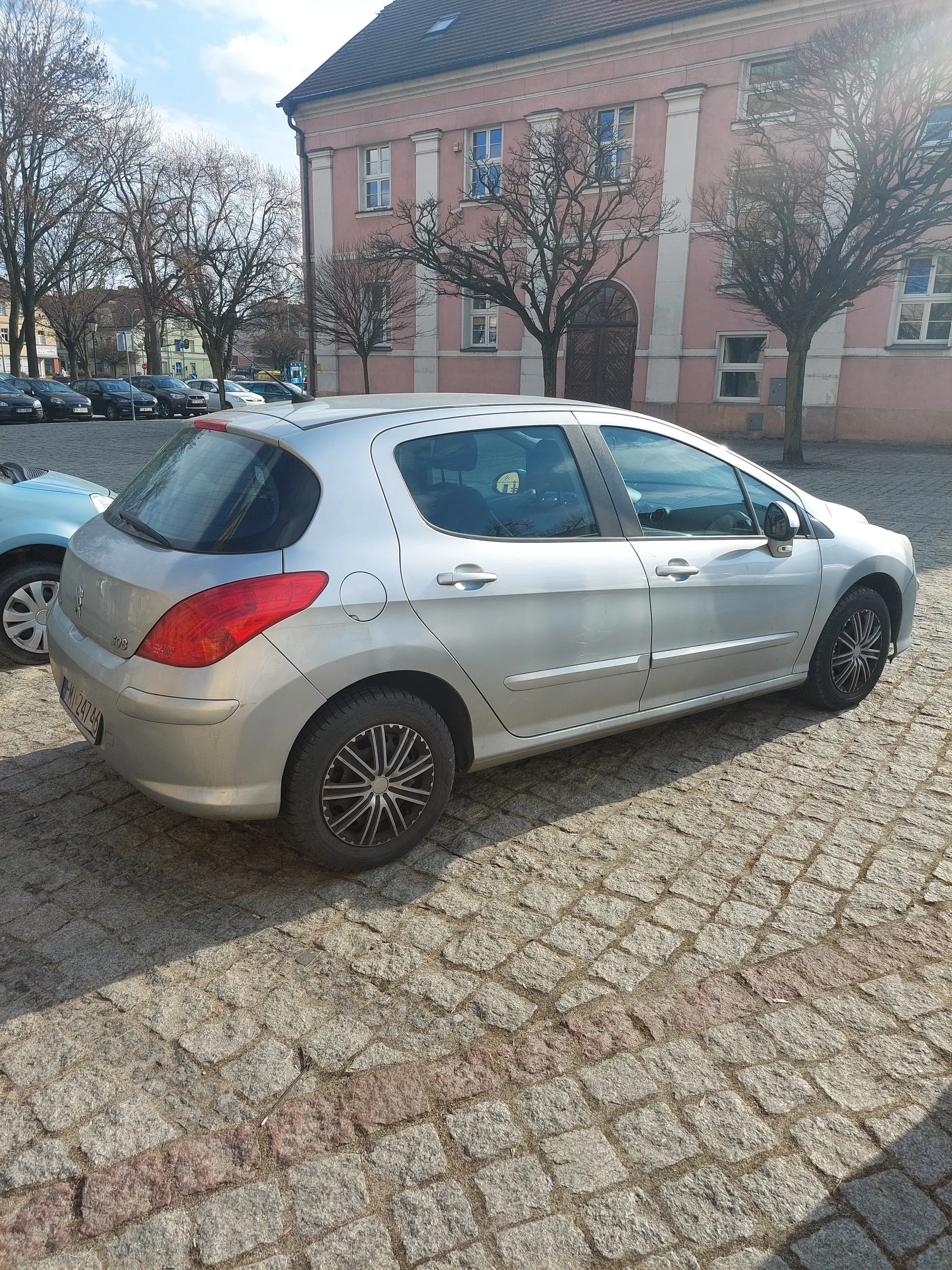 Srebrny samochód osobowy zaparkowany na brukowanej ulicy. W tle różowy budynek z białymi elementami, bezlistne drzewa oraz inne zaparkowane pojazdy. Niebo częściowo zachmurzone.