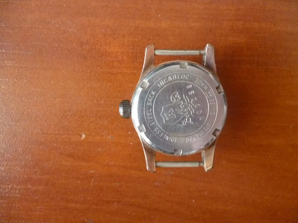 Zdjęcie przedstawiające tył srebrnego zegarka na brązowym, drewnianym tle. Na metalowej kopercie widnieją grawery: 'STAINLESS STEEL BACK', 'INCABLOC', 'SWISS MADE', 'WATERPROOF' oraz logo z napisem 'Delux'. Widoczny jest również numer seryjny '207259'. Zegarek nie posiada paska.