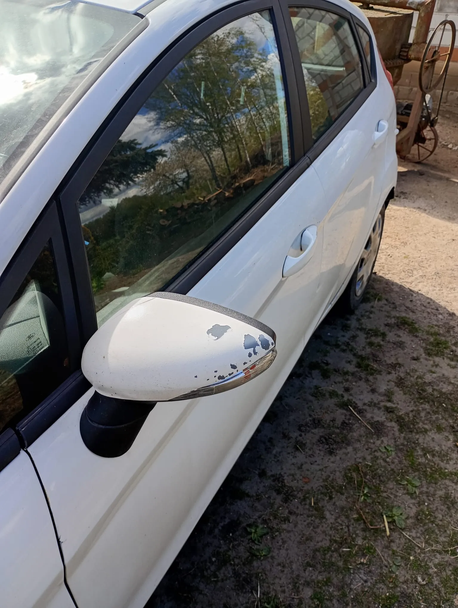 Biały samochód widziany z boku, z uszkodzonym lusterkiem bocznym (zadrapana farba). Samochód zaparkowany na podłożu z ziemią i trawą. W tle widoczne stare, rdzewiałe maszyny rolnicze oraz drzewa odbijające się w szybach pojazdu. Niebo częściowo zachmurzone.