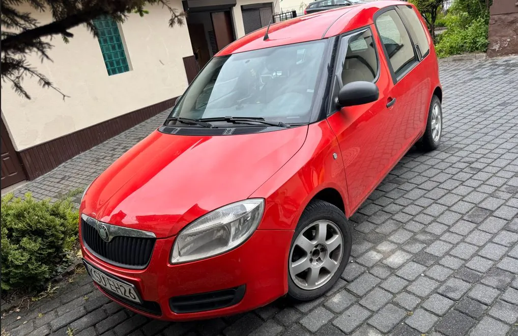 Zdjęcie przedstawia czerwony samochód marki Skoda zaparkowany na brukowanej nawierzchni. Widoczna tablica rejestracyjna z numerem G07H26. W tle budynek z beżową ścianą i zielonym oknem, krzewy oraz gałąź drzewa. Samochód ma srebrne felgi i czarne lusterka boczne.