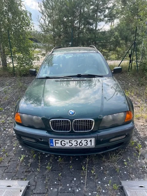 Zdjęcie przodu ciemnozielonego samochodu marki BMW zaparkowanego na kostce brukowej. Samochód ma polską tablicę rejestracyjną o numerze FG 53631. W tle widoczne są drzewa i zielony płot z siatki.