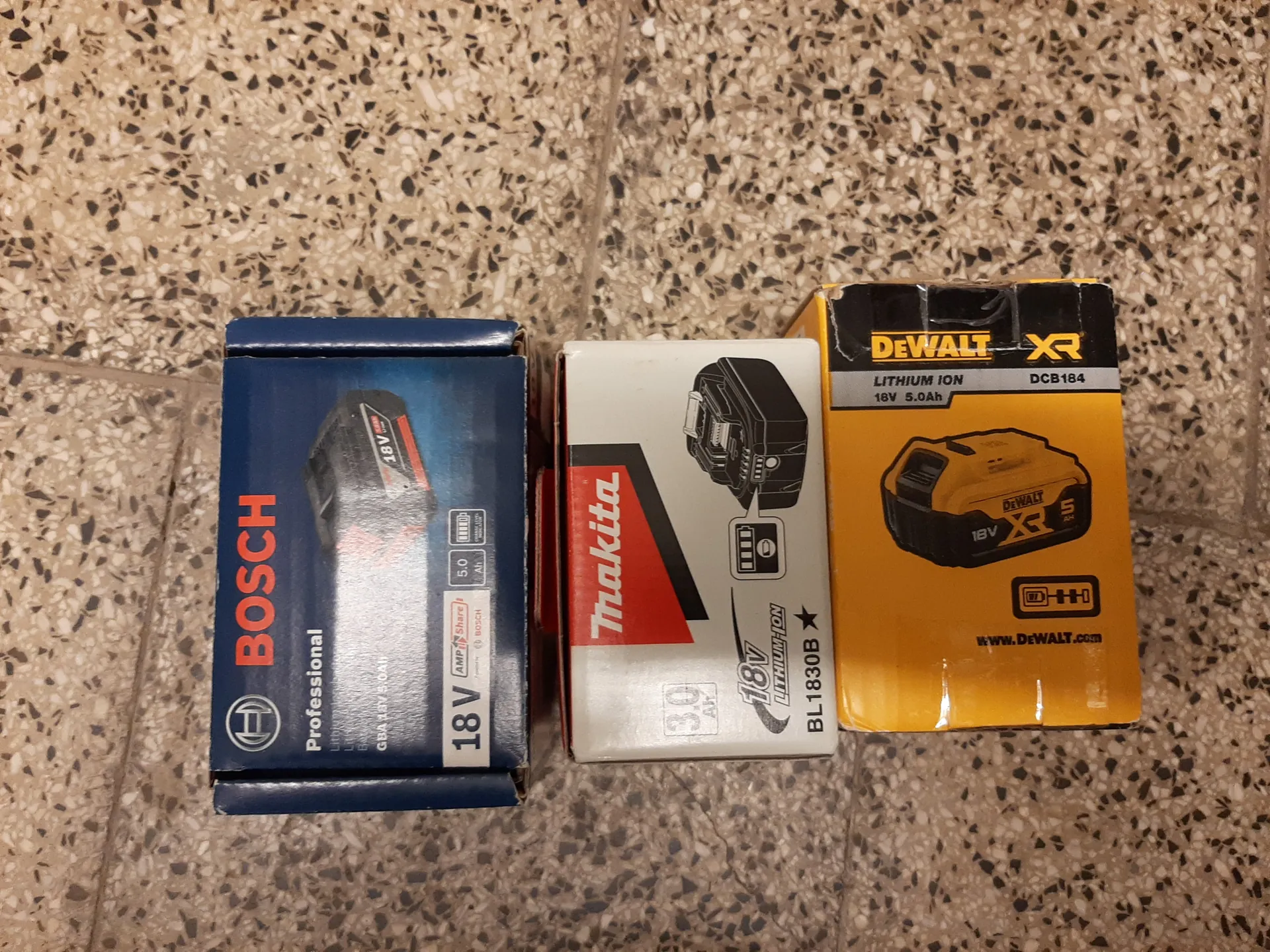 Trzy pionowo ułożone pudełka z akumulatorami na podłodze z kruszywem. Na górze niebieskie pudełko marki BOSCH Professional z napisem 18V, 5.0 Ah i obrazem akumulatora. W środku białe pudełko marki Makita z czerwonym pasem, napisem 18V LITHIUM-ION, 3.0 Ah i modelem BL1830B. Na dole żółte pudełko marki DEWALT XR z czarnym pasem, napisem LITHIUM ION 18V 5.0Ah i modelem DCB184. Tekst na wszystkich pudełkach jest czytelny i ustawiony pionowo.