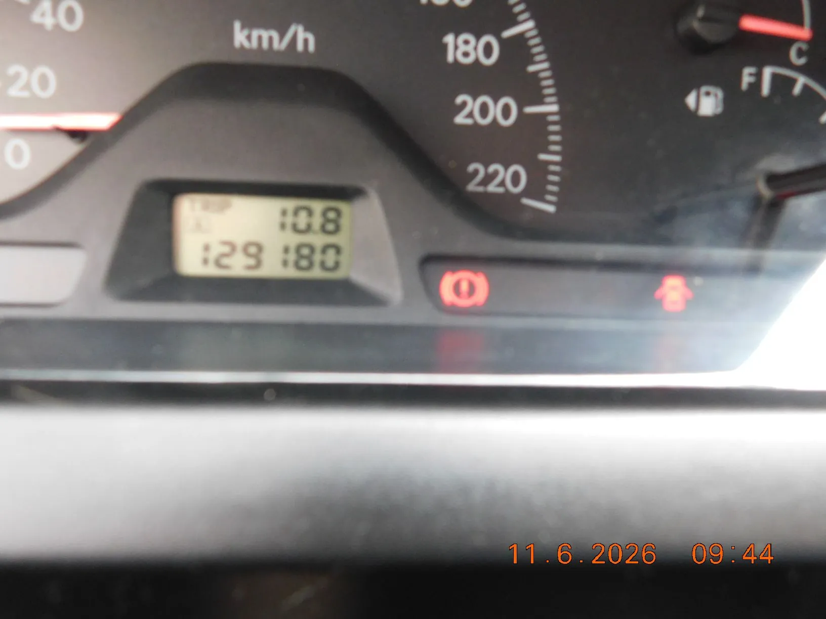 Zbliżenie na deskę rozdzielczą samochodu. Widoczny jest fragment prędkościomierza z oznaczeniami 180, 200 i 220 km/h. Mały cyfrowy wyświetlacz pokazuje wartości 10.8 oraz 129 180. Poniżej znajdują się dwie czerwone kontrolki ostrzegawcze. W prawym dolnym rogu zdjęcia widnieje pomarańczowa data i godzina: 11.6.2026 09:44.