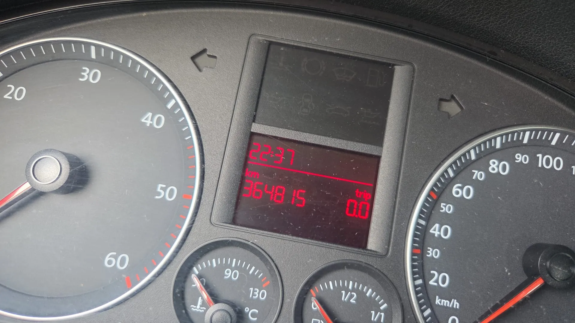 Zbliżenie na deski rozdzielcze samochodu. Po lewej stronie znajduje się wskaźnik obrotomieru z czerwonym wskazówką, skala od 0 do 60. W centrum wyświetlacz cyfrowy z czerwonymi cyframi: godzina 22:37, przebieg 364815 km oraz trip 0.0. Po prawej stronie wskaźnik prędkości w km/h z zakresem do 100. Poniżej znajdują się dwa mniejsze wskaźniki: termometr z zakresem do 130°C i wskaźnik paliwa z podziałką 1/2 i 1/1. Na górze wyświetlacza widoczne są symbole ostrzegawcze. Wszystkie elementy są wyraźnie widoczne, a tekst na wyświetlaczu jest czytelny.