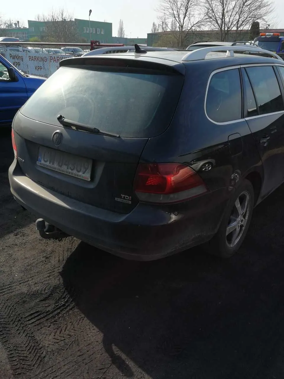 Czarny samochód osobowy typu kombi marki Volkswagen z widocznym tyłem, tablicą rejestracyjną CW 04309 oraz napisem TDI na bagażniku. Po lewej stronie niebieski samochód, w tle biały płot z napisami Parking KPP, budynki (w tym zielony z napisem DODA) oraz drzewa bez liści. Samochód stoi na asfaltowej powierzchni z widocznymi śladami opon.