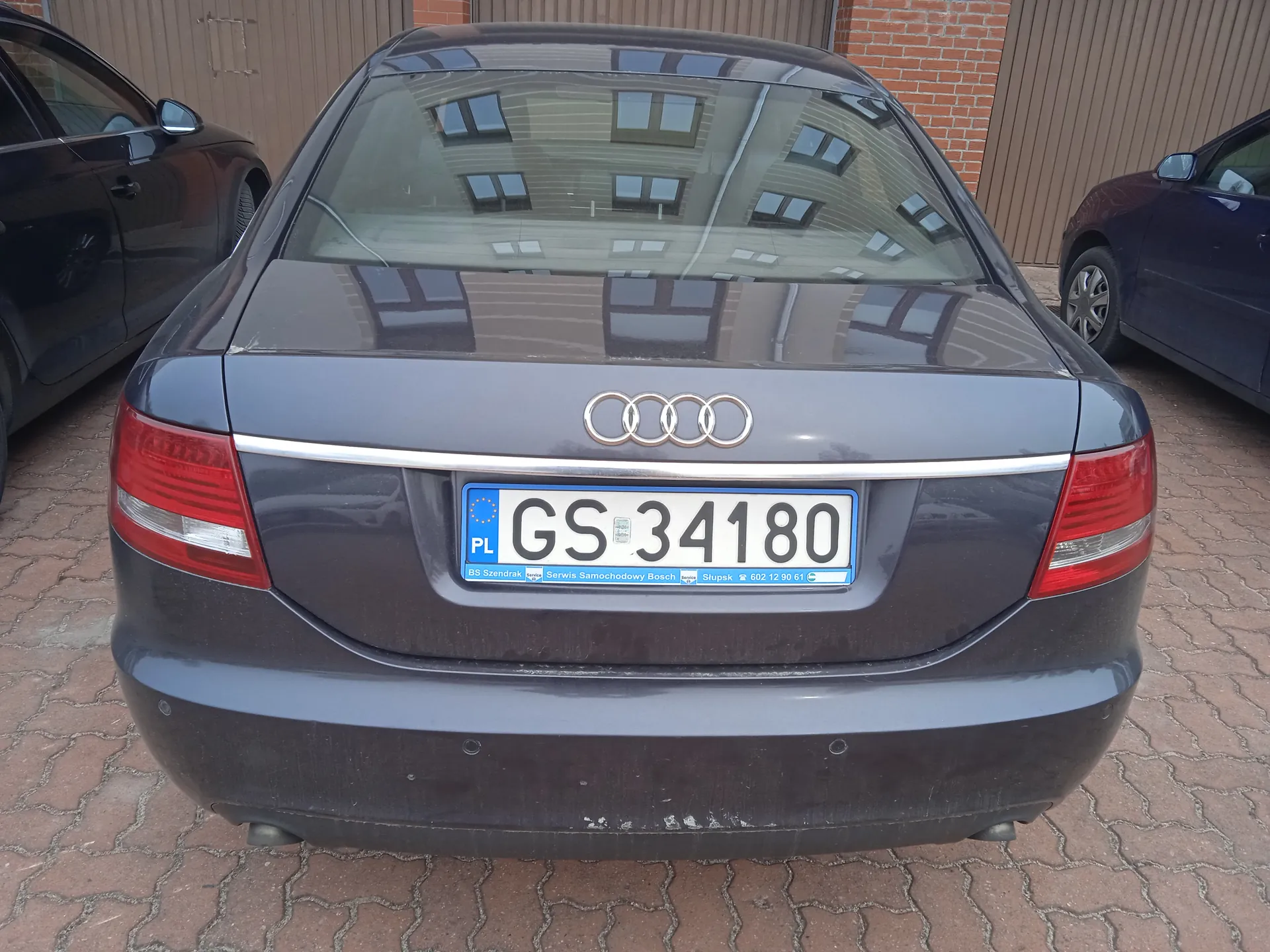 Tył szarego samochodu marki Audi z widocznym logo czterech pierścieni. Tablica rejestracyjna z białym tłem i czarnym napisem GS 34180, po lewej stronie niebieski pas z napisem PL oraz niebieską taśmą zawierającą informacje o serwisie. Samochód zaparkowany na brukowanej powierzchni, po obu stronach widoczne fragmenty innych pojazdów (czarny po lewej, ciemnoniebieski po prawej). W tle budynki z brązowymi panelami i ceglaną ścianą. Odbicie budynku w tylnej szybie samochodu.
