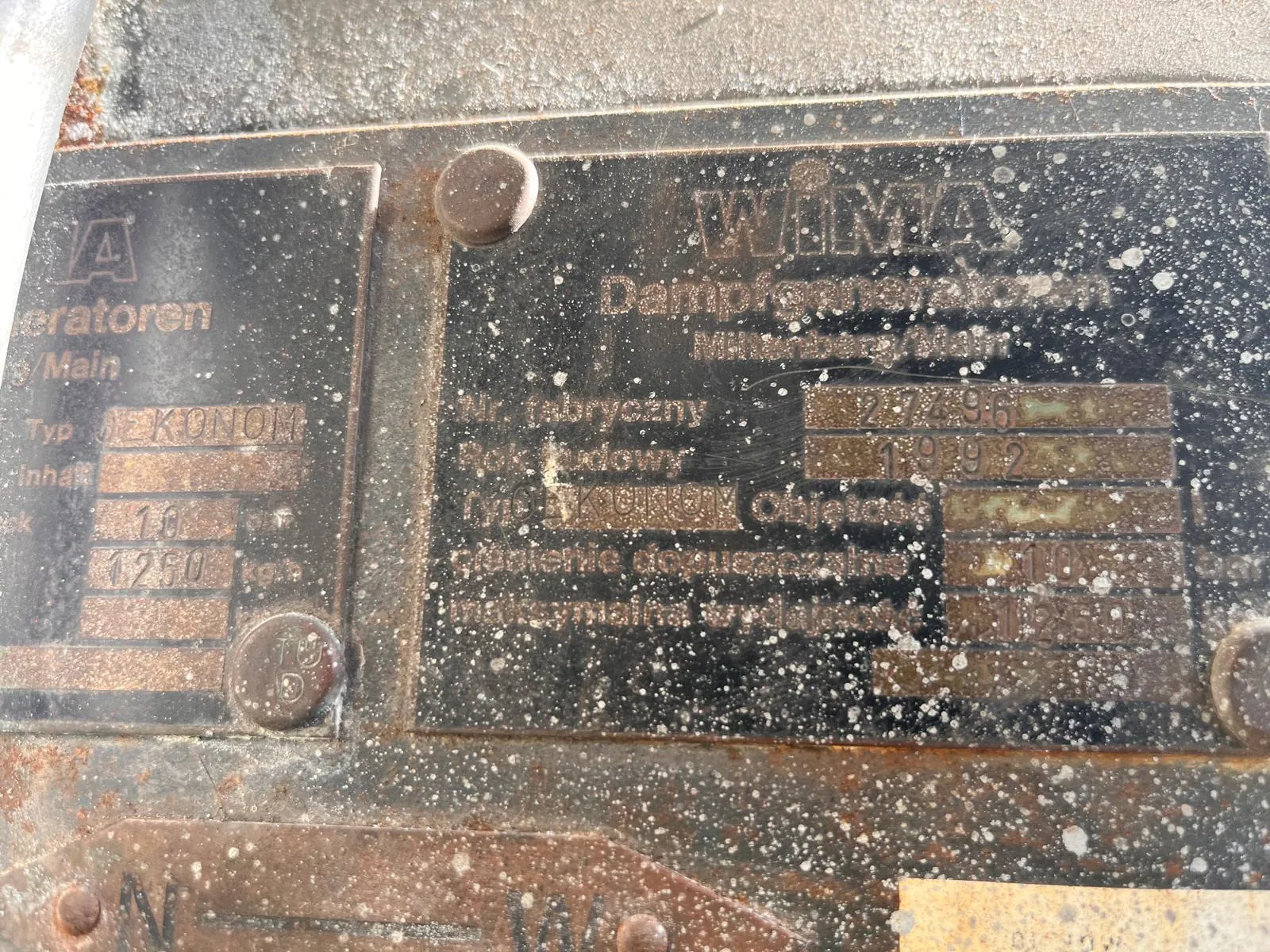 Zbliżenie na dwie metalowe tabliczki znamionowe przymocowane do zardzewiałej, metalowej powierzchni. Tabliczki są czarne z wytłoczonymi i wyrytymi napisami w języku niemieckim. Na większej tabliczce widnieje napis 'WIMA' oraz 'Dampfpressmaschine'. Poniżej znajdują się pola z danymi: 'Nr. fabryczny', 'Rok budowy' (widoczna data 1929), 'Typ' oraz inne parametry techniczne. Mniejsza tabliczka po lewej zawiera napis 'eratoren' oraz 'Typ EKONOM'. Powierzchnia metalu jest pokryta rdzą i białymi odpryskami farby lub osadem.