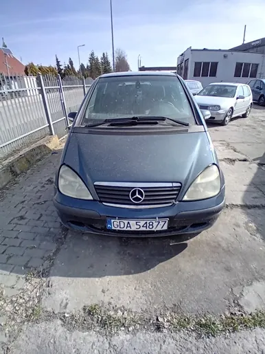 Ciemnoszary samochód marki Mercedes-Benz z tablicą rejestracyjną GDA 54877, zaparkowany na betonowej powierzchni. W tle metalowe ogrodzenie, inne zaparkowane pojazdy (w tym biały samochód), budynki przemysłowe oraz drzewa. Niebo jest jasne z nielicznymi chmurami. Na pierwszym planie widoczne są fragmenty brukowanej nawierzchni i trawy.