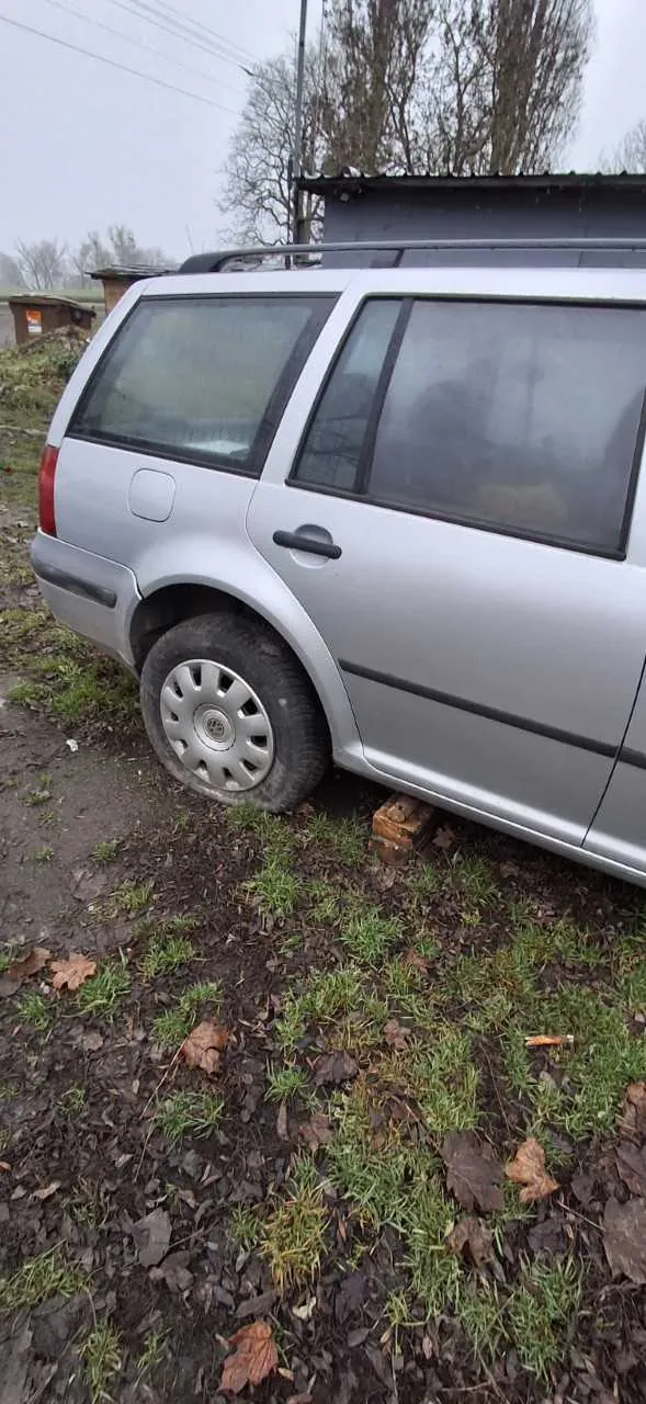 Srebrny samochód typu kombi zaparkowany na trawiastym i błotnistym terenie z rozsypanymi liśćmi. Tylna opona opiera się na cegle. W tle widoczne są drzewa bez liści, niewielki budynek z metalowym dachem oraz słupy energetyczne. Niebo jest zachmurzone. Brak czytelnego tekstu.