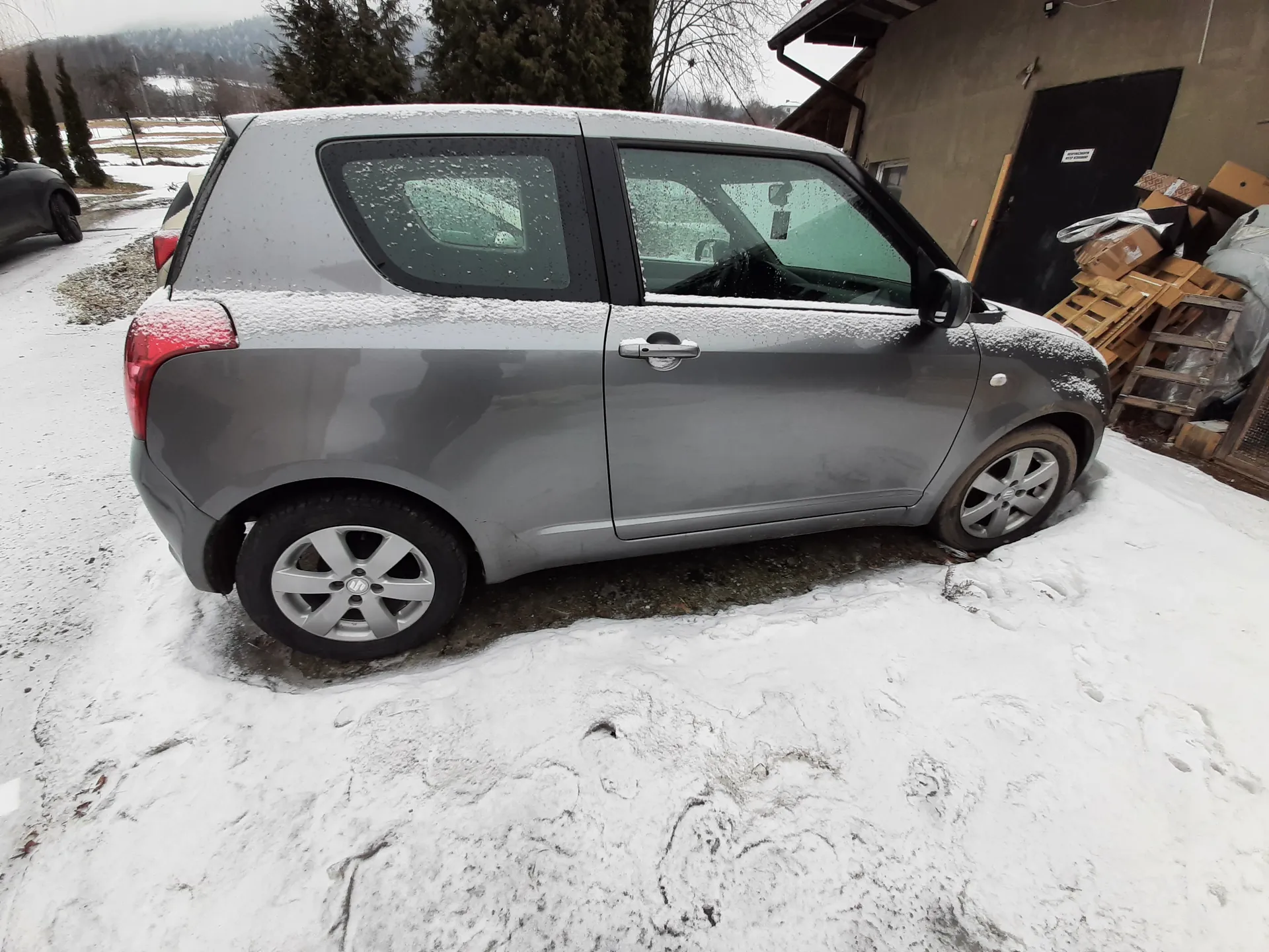 Szary dwudrzwiowy samochód pokryty śniegiem na dachu i szybach, zaparkowany na śnieżnej nawierzchni obok budynku z czarnym wejściem, paletami drewnianymi i kartonami. W tle drzewa i zimowy krajobraz.