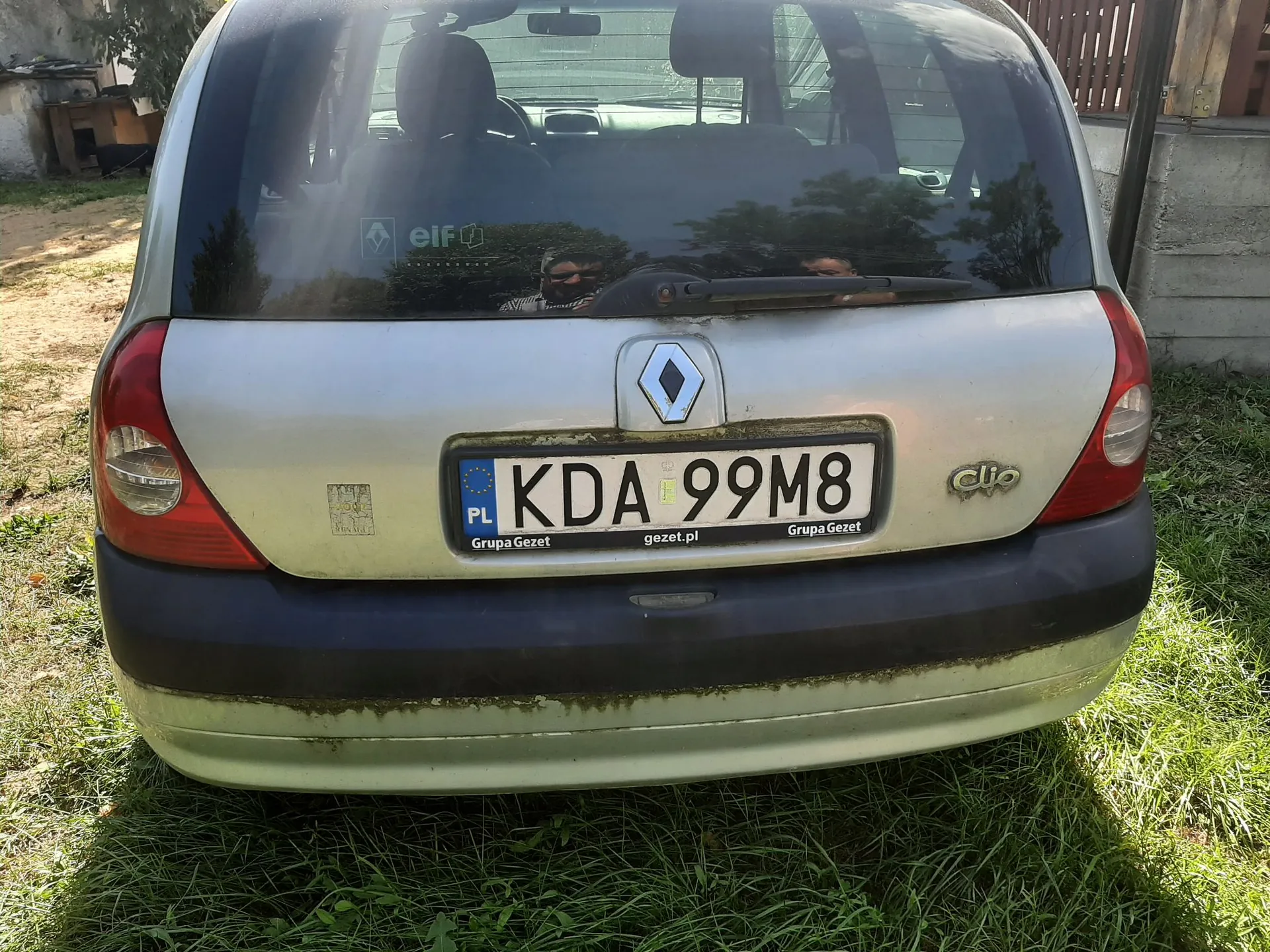 Tylny widok srebrnego samochodu marki Renault Clio zaparkowanego na trawie. Na klapie bagażnika znajduje się logo Renault oraz polska tablica rejestracyjna o numerze KDA 99M8. Poniżej tablicy widnieje napis 'Grupa Gezet' oraz adres strony internetowej 'gezet.pl'. Samochód ma ciemny, plastikowy zderzak z widocznymi zabrudzeniami.