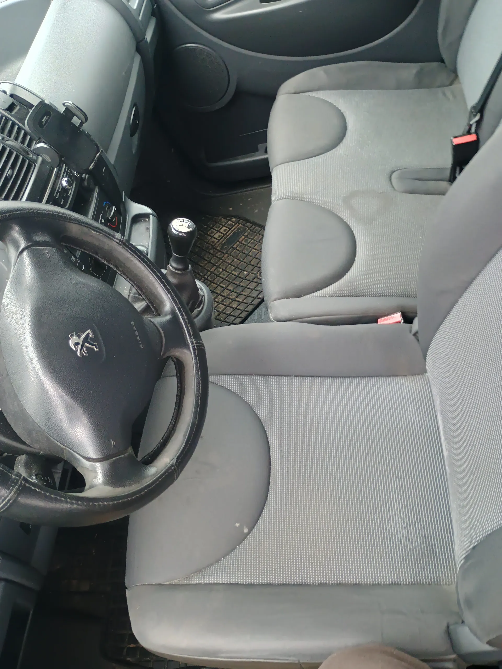 Wnętrze samochodu z kierownicą z logo Peugeot, szarymi siedzeniami tapicerowanymi tkaniną, dźwignią zmiany biegów, deską rozdzielczą z przyciskami i dywanikami podłogowymi.