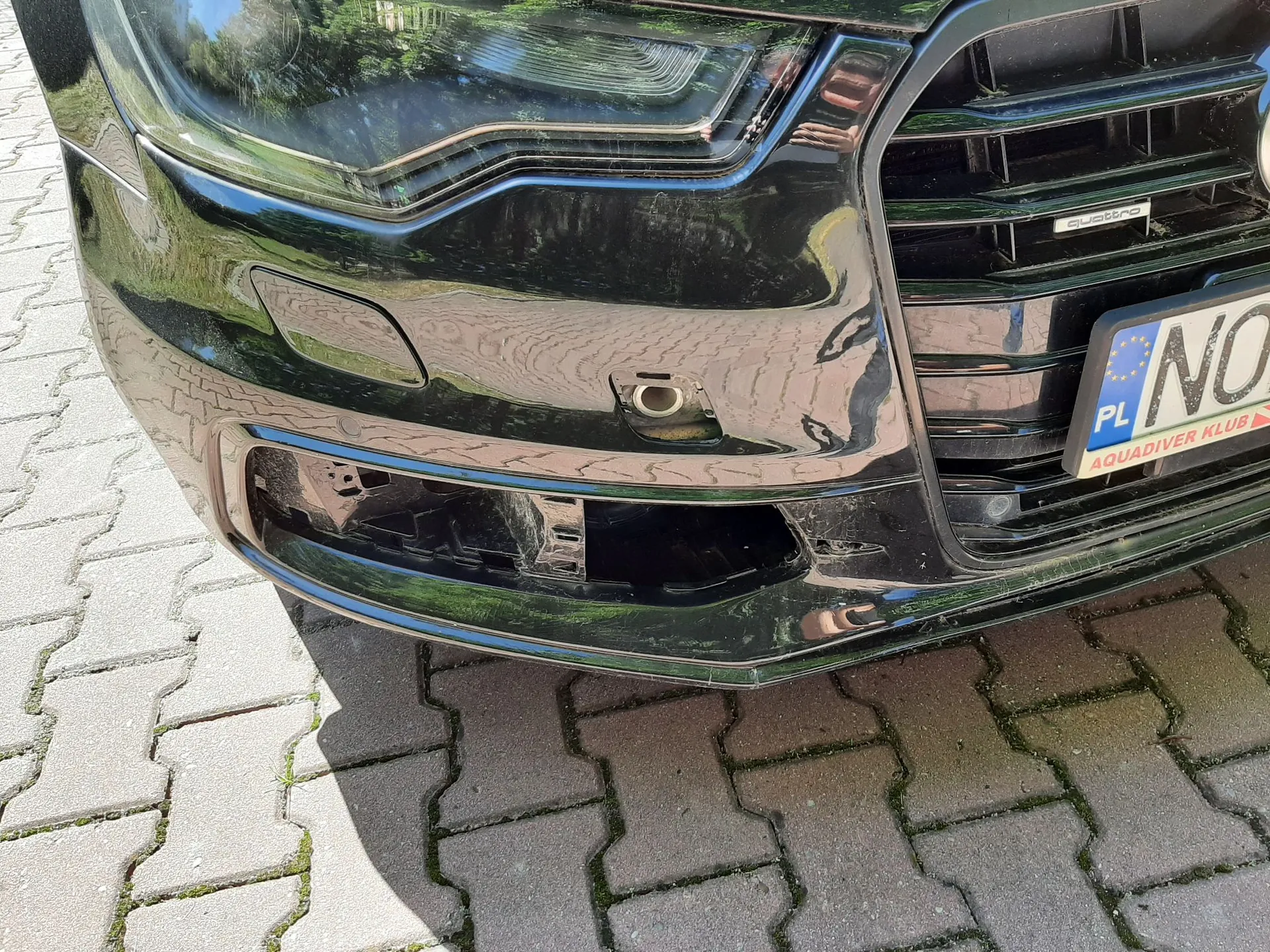 Zbliżenie na przedni czarny zderzak samochodu marki Audi. Widoczny jest fragment reflektora, grill z napisem 'quattro' oraz fragment tablicy rejestracyjnej z oznaczeniem 'PL' i napisem 'AQUADIVER KLUB' na ramce. W dolnej części zderzaka znajduje się ubytek w plastikowym elemencie wykończenia oraz widoczny otwór montażowy. Samochód stoi na szarym bruku z kostki betonowej.