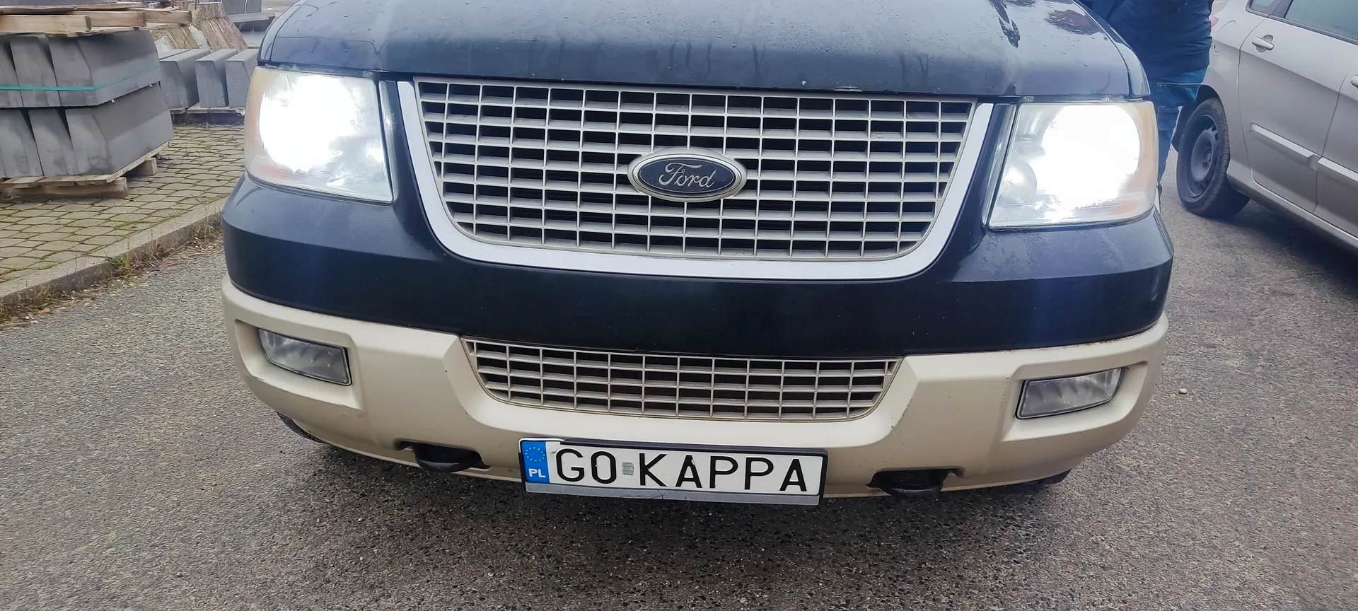 Przednia część ciemnoniebieskiego samochodu marki Ford z beżowym zderzakiem. Na środku atrapy chłodnicy znajduje się logo Ford. Poniżej tablica rejestracyjna z napisem GO KAPPA oraz oznaczeniem PL po lewej stronie. Po prawej stronie częściowo widoczny srebrny samochód, po lewej stosy betonowych elementów na podkładach. Tło stanowi asfaltowa powierzchnia i brukowana droga.