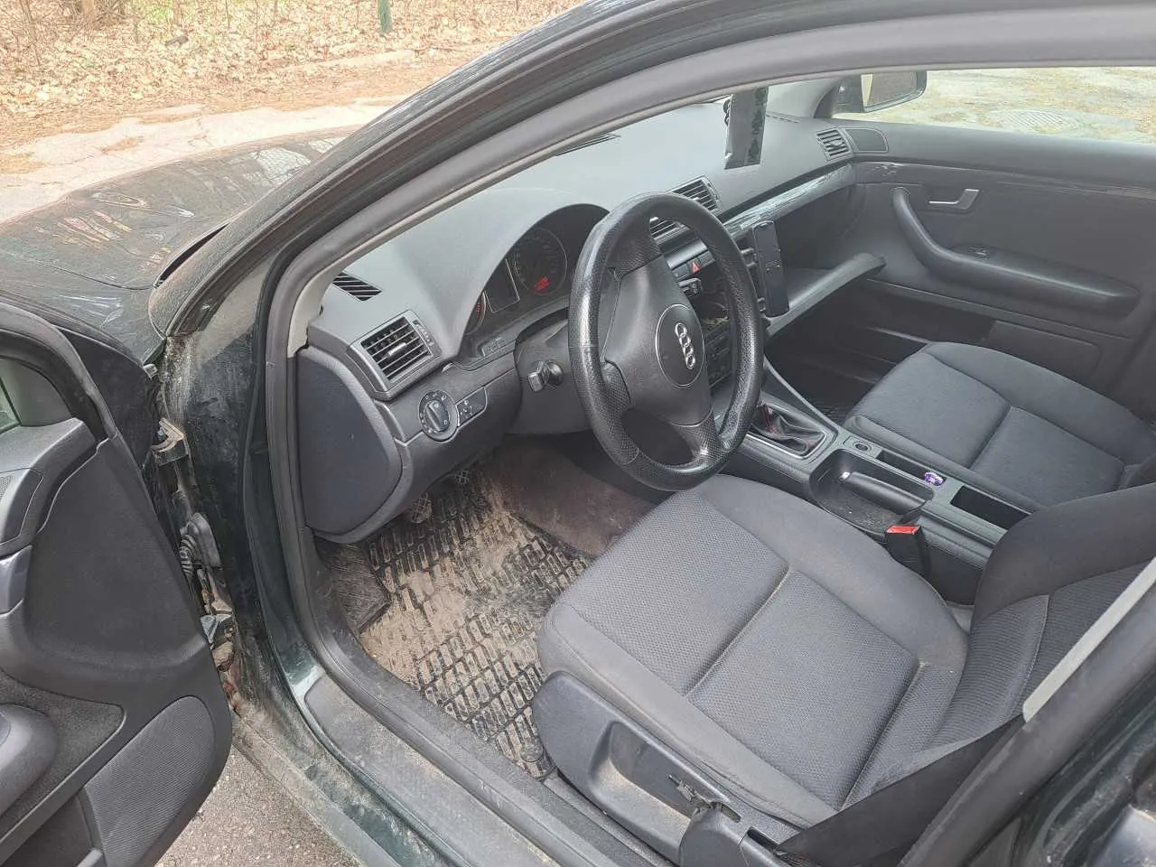 Wnętrze czarnego samochodu marki Audi z otwartą lewą przodem drzwiami. Widoczne są czarne siedzenia tapicerowane tkaniną, kierownica z logo Audi, deska rozdzielcza z licznikami, klimatyzacją i przyciskami sterującymi. Na podłodze znajdują się brudne dywaniki z widocznymi śladami ziemi i liści. Poza samochodem widoczna jest droga oraz obszar z suchymi liśćmi na ziemi.