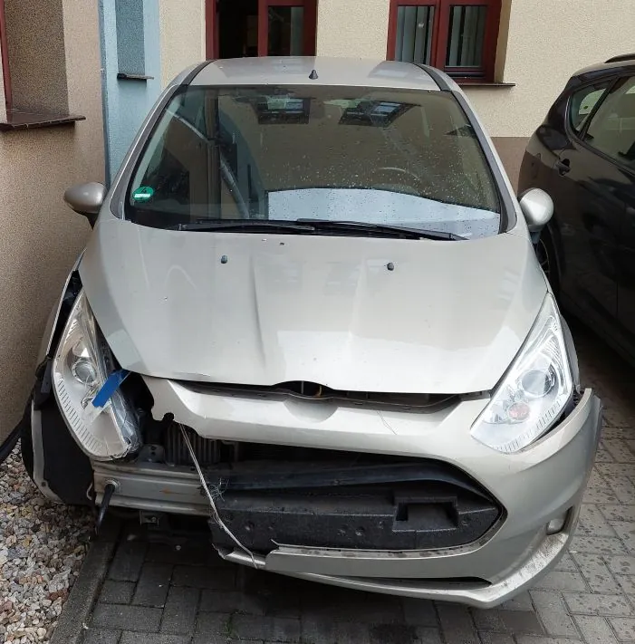 Srebrny samochód osobowy widziany od przodu, zaparkowany na kostce brukowej przed budynkiem. Pojazd ma poważne uszkodzenia przedniego zderzaka, który jest częściowo oderwany i zapadnięty do wewnątrz. Lewa reflektor jest poluzowany i podtrzymywany niebieską taśmą. Maska samochodu jest lekko uchylona.