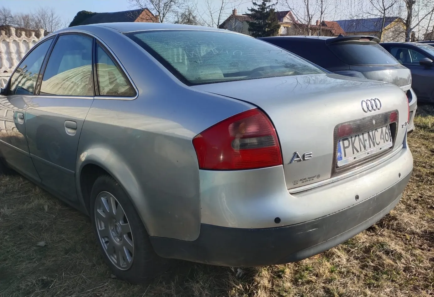 Srebrny samochód Audi A6 widziany od tyłu, z tablicą rejestracyjną PKN NC 46. Pojazd stoi na trawie, w tle widoczne są inne samochody, domy i drzewa bez liści. Na bagażniku umieszczono logo Audi oraz oznaczenie A6. Tło stanowi jasne niebo i budynki mieszkalne.