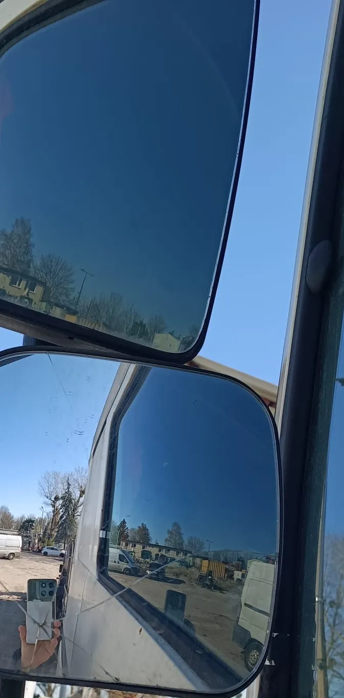 Zdjęcie wykonane z wnętrza pojazdu, pokazujące dwa lusterka boczne. W górnym lusterku odbija się bezchmurne, niebieskie niebo oraz wierzchołki drzew i budynków. W dolnym, większym lusterku widoczne jest odbicie białego vana, placu budowy z maszynami, innych pojazdów oraz osoby robiącej zdjęcie telefonem komórkowym.