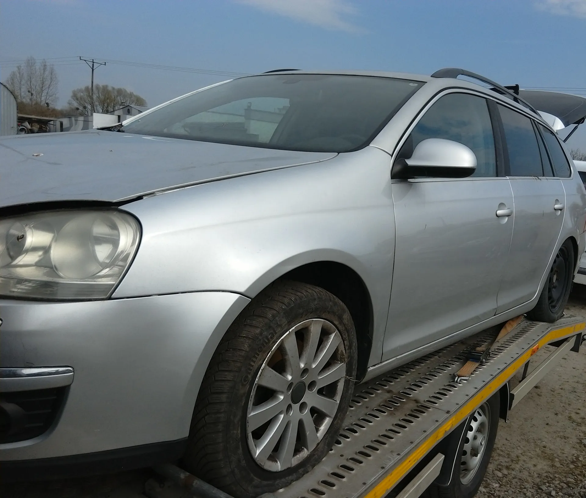 Srebrny samochód typu kombi z otwartą maską i uszkodzonym przednim zderzakiem, umieszczony na płycie holownika. Widoczne są felgi, dachowy bagażnik oraz tło z drzewami, słupami energetycznymi i budynkami pod jasnym niebem.