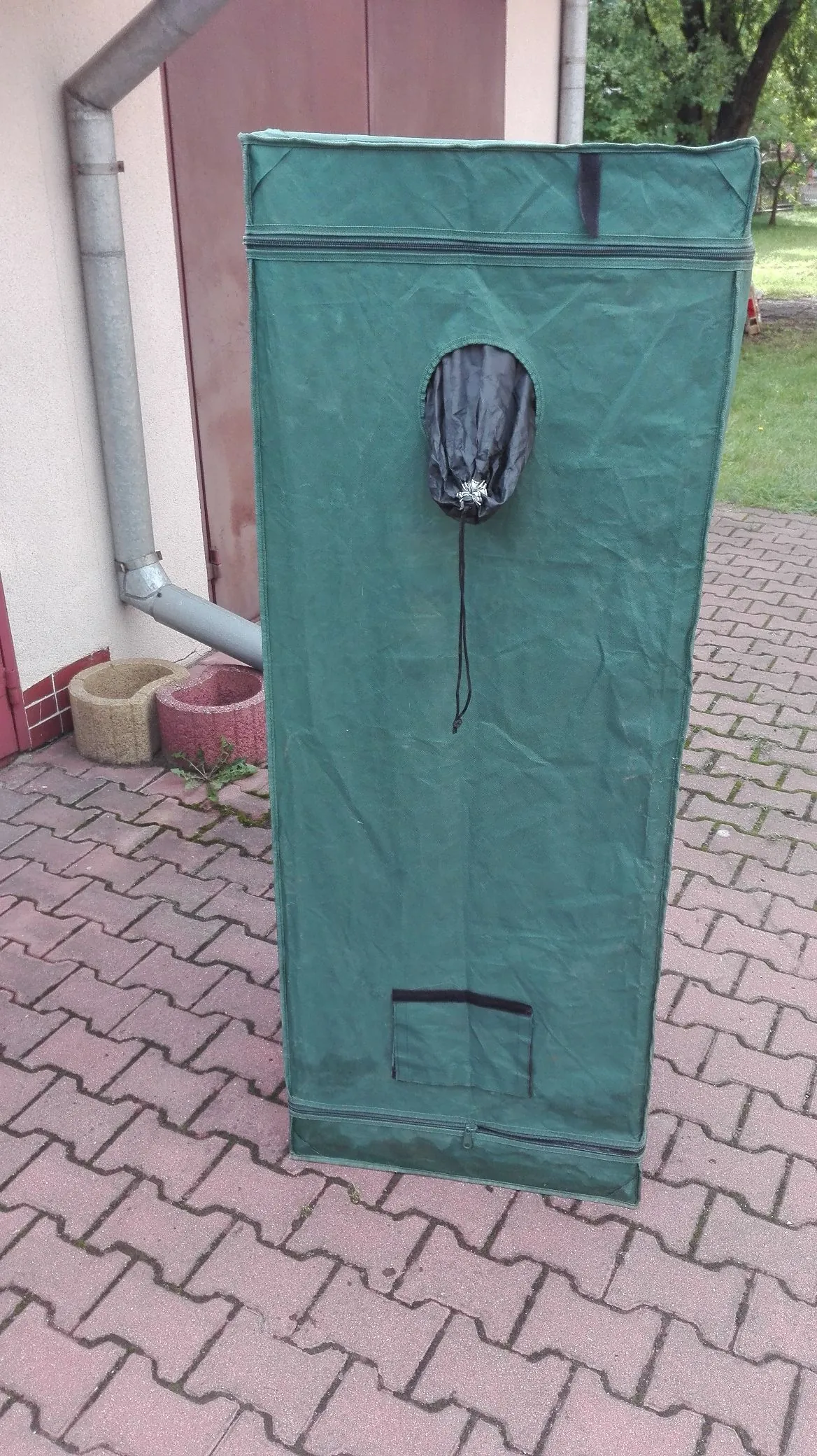 Zielona, materiałowa szafa przenośna stojąca na kostce brukowej przed budynkiem. Szafa ma okrągły otwór z czarną, ściąganą tkaniną w górnej części oraz małą kieszeń na dole. W tle widoczna jest ściana budynku, rura spustowa oraz dwie donice.