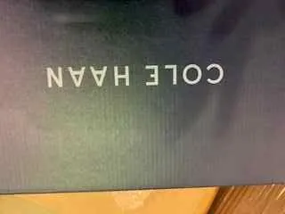 Ciemnoniebieskie tło z białym, pionowym napisem 'COLE HAAN'. Po lewej stronie widoczna jest drewniana powierzchnia.