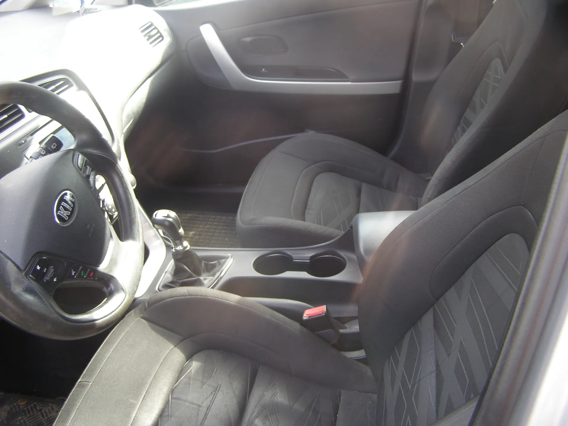 Wnętrze samochodu marki Kia z widocznym kierownicą z logo Kia, czarnymi siedzeniami z geometrycznym wzorem, dźwignią zmiany biegów, dwoma uchwytami na napoje oraz fragmentem deski rozdzielczej i drzwi pasażera. Wnętrze posiada czarne i srebrne elementy wykończenia.