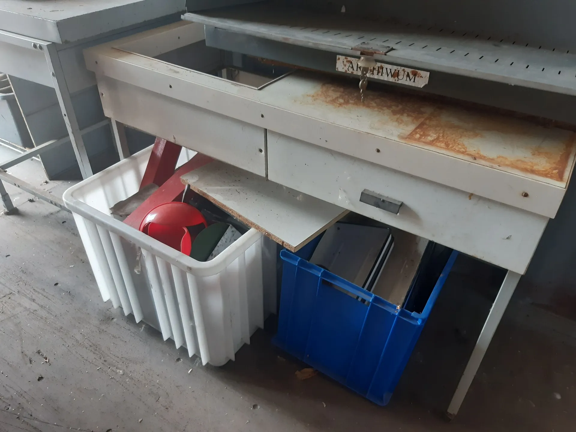 Zdjęcie przedstawiające biały, metalowy stół warsztatowy z wysuniętą szufladą, która jest zardzewiała na wierzchu. Pod stołem znajdują się dwa plastikowe pojemniki: biały kosz z białymi pionowymi żłobieniami, w którym widać czerwone i zielone przedmioty, oraz niebieska skrzynka plastikowa zawierająca szare teczki lub foldery. Na krawędzi blatu widoczna jest mała, zardzewiała tabliczka z napisem 'ALUMIUM'.