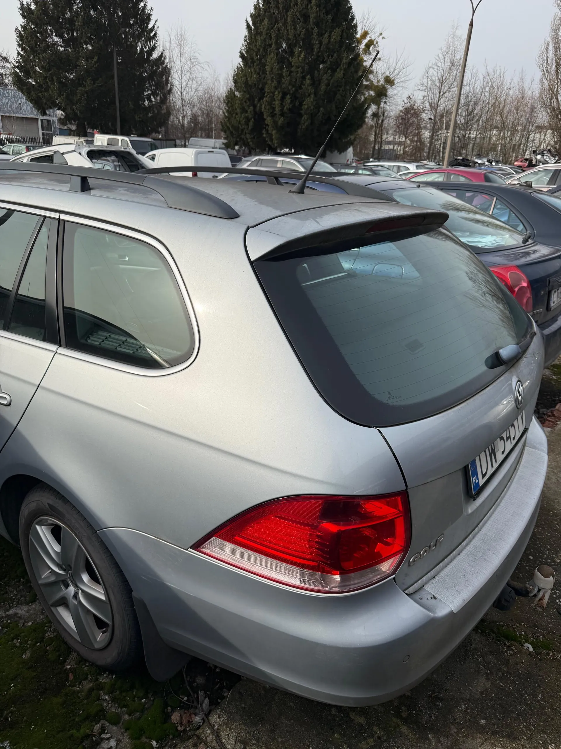 Srebrny samochód osobowy typu kombi Volkswagen Golf z widocznym tyłem, czerwonymi światłami tylnymi i tablicą rejestracyjną PL DW 5451V. Samochód stoi na otwartym parkingu wśród innych pojazdów. W tle drzewa, budynki i słup latarni. Podwozie częściowo pokryte mchem, a na ziemi widoczne są kamienie i sucha trawa.