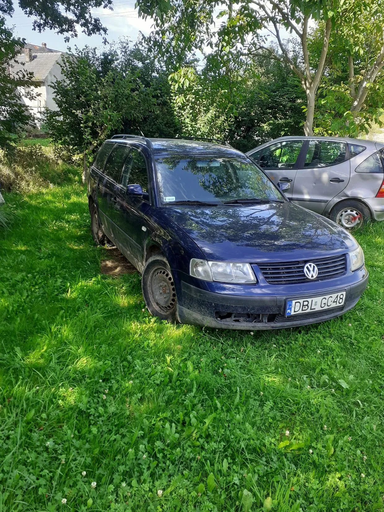 Ciemnoniebieski samochód osobowy marki Volkswagen w nadwoziu kombi, zaparkowany na zielonej trawie. Pojazd ma polską tablicę rejestracyjną o numerze DBL GC48. W tle widoczny jest srebrny samochód, gęste zielone krzewy, drzewa oraz fragment białego budynku z dachem.