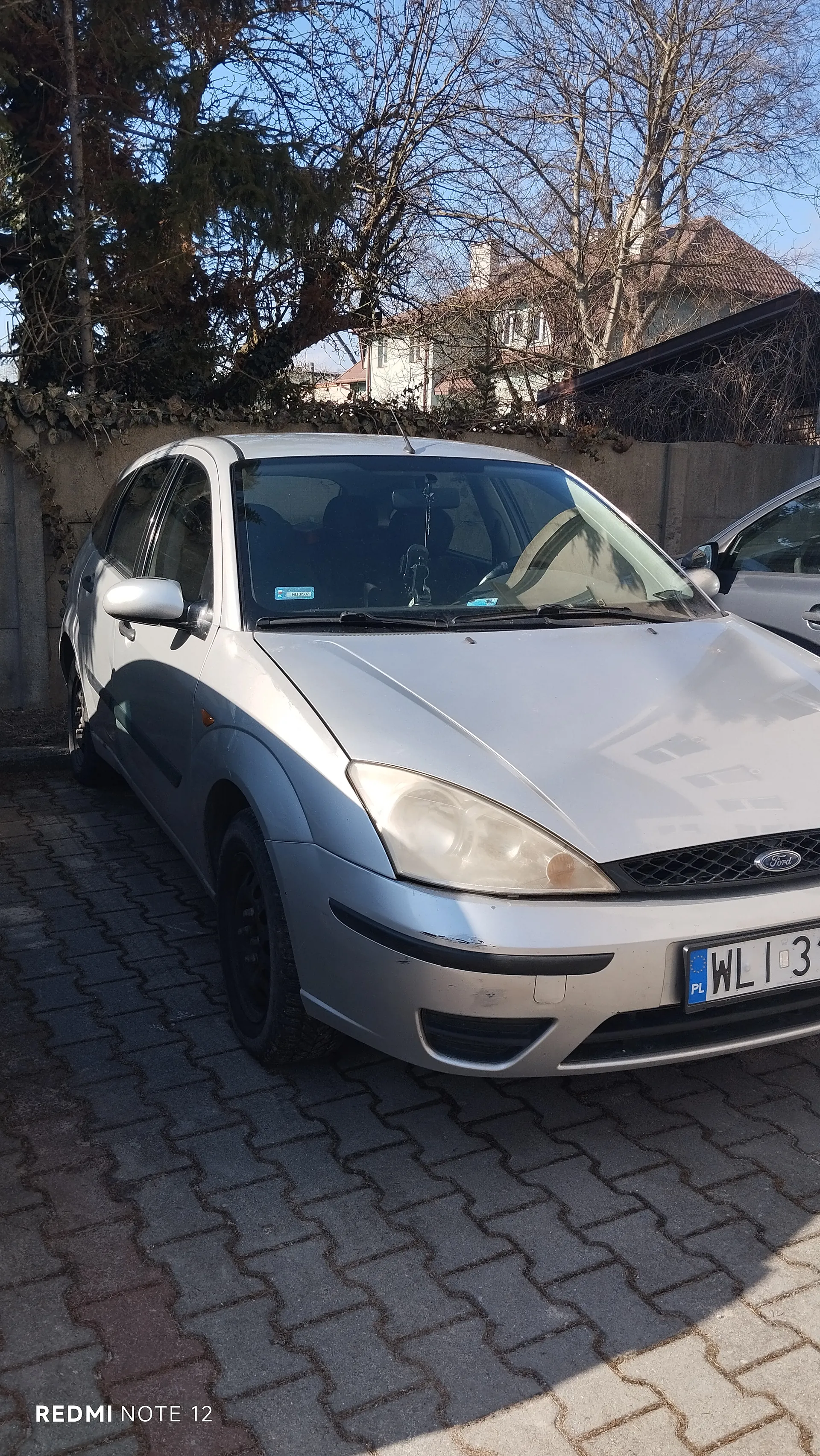 Srebrny samochód marki Ford zaparkowany na brukowanej powierzchni. Widoczna tablica rejestracyjna z napisem PL WL1 3 oraz logo Ford na atrapie chłodnicy. W tle betonowy mur porośnięty pnączami, drzewa bez liści, domy i jasne niebo. Po prawej stronie częściowo widoczny inny samochód. Na dole zdjęcia napis REDMI NOTE 12.
