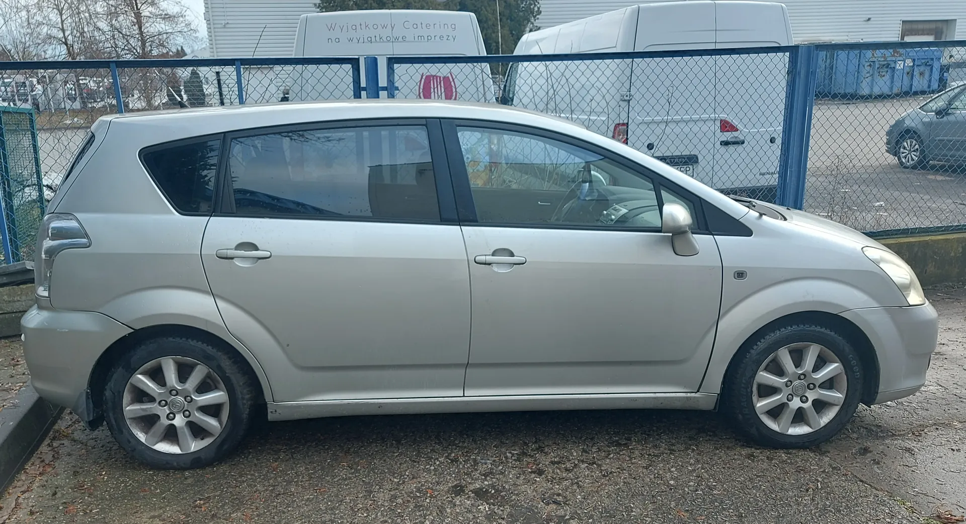 Srebrny samochód osobowy typu minivan zaparkowany na podłożu z drobnej kostki. Po lewej stronie widoczna czarna krawężnik, za samochodem siatkowy płot z niebieskimi słupkami. Za płotem dwa białe furgony, na jednym z nich napis: "Wyjątkowy Catering na wyjątkowe imprezy" oraz różowy logo. W tle budynki, suche drzewa i szary samochód zaparkowany na asfalcie. Podłoże wokół samochodu pokryte suchymi liśćmi i gruzem.