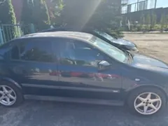 Profil ciemnoniebieskiego samochodu osobowego zaparkowanego na utwardzonym placu. W tle widoczne są zielone drzewa, krzewy oraz fragmenty budynków pod jasnym niebem z silnym odblaskiem słońca.