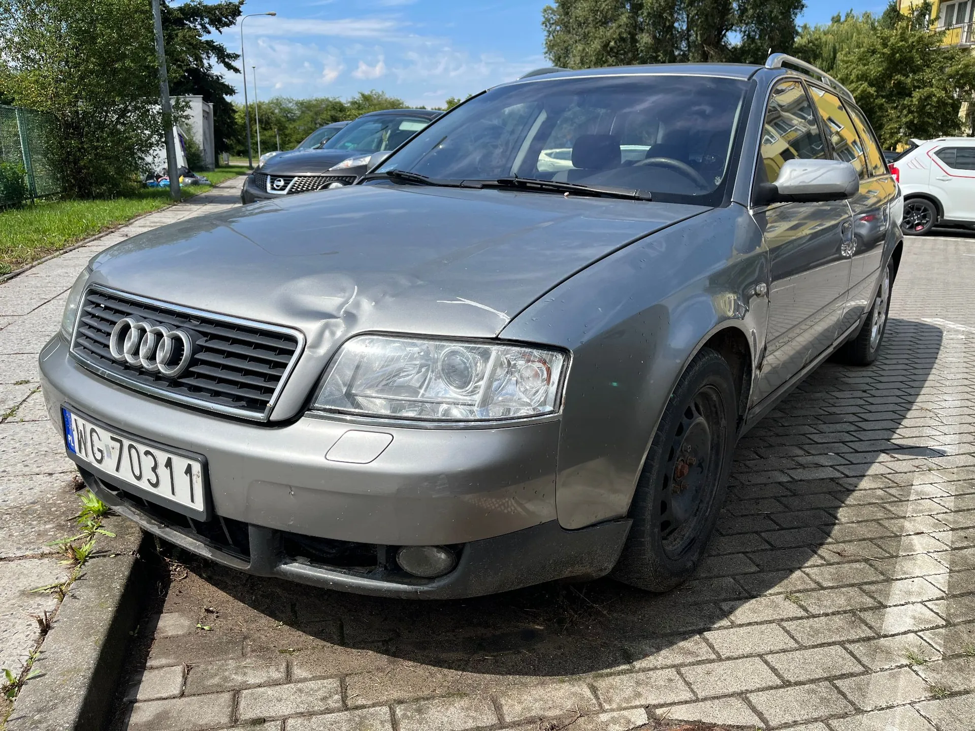 Srebrnoszary samochód marki Audi w nadwoziu kombi, zaparkowany na kostce brukowej. Pojazd ma widoczne uszkodzenia karoserii: wgniecenie na masce nad lewym reflektorem oraz wgniecenie na przednim lewym błotniku. Samochód posiada polską tablicę rejestracyjną o numerze WG 70311. W tle widoczny jest inny ciemny samochód, zieleń oraz fragmenty budynków pod błękitnym niebem.